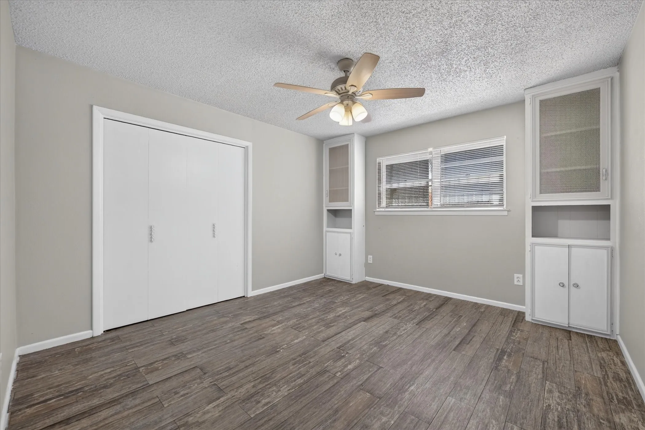 Wood-finish flooring, ceiling fan, white bi-fold closet doors, built-in cabinetry with glass-front display, and a window with horizontal blinds