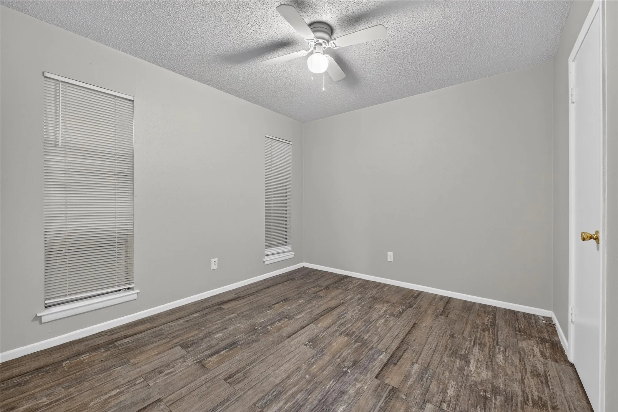 Wood-finish flooring, two windows with blinds, ceiling fan, and light gray wall paint