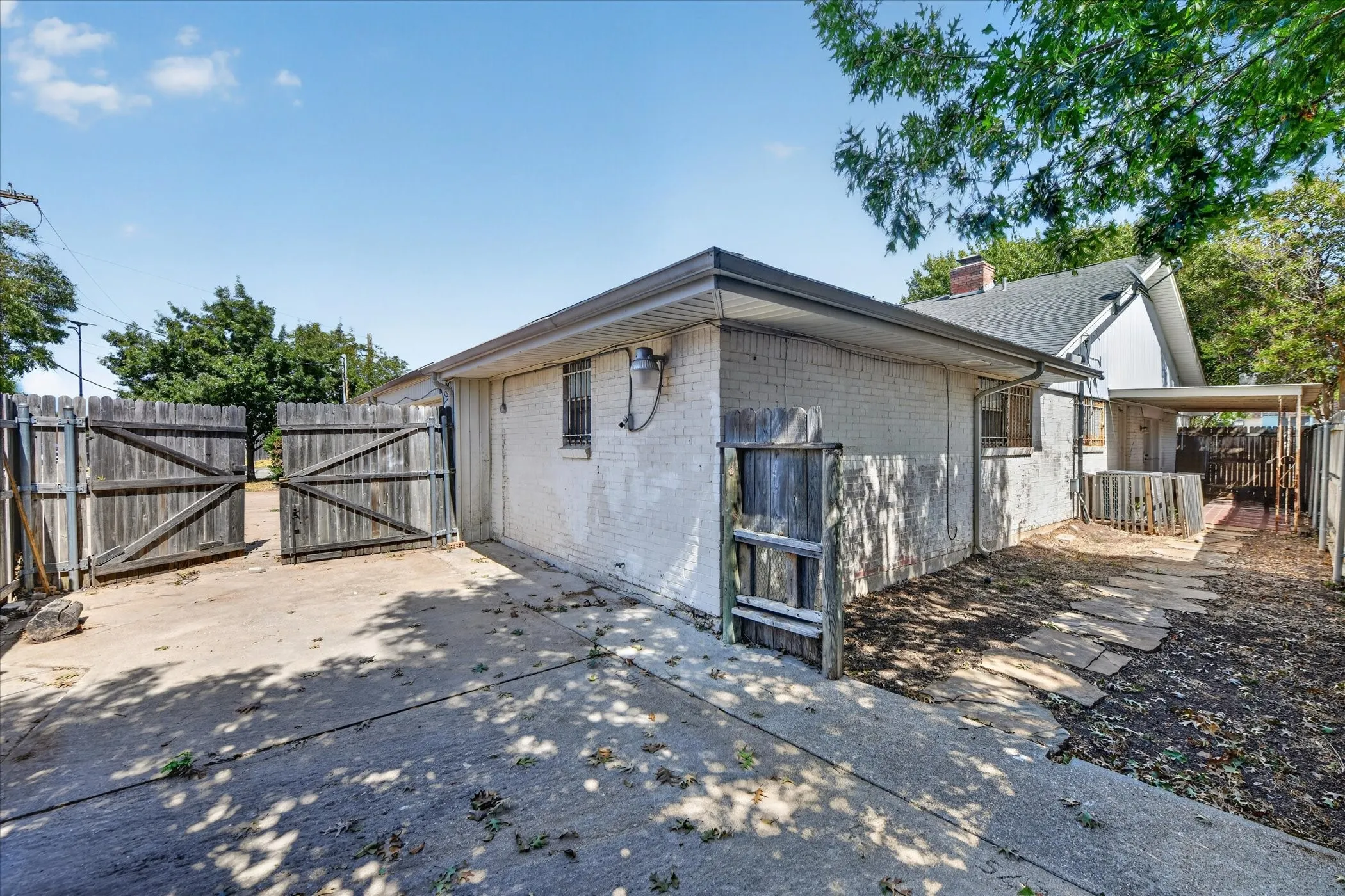 Concrete driveway with double wood gates, textured exterior walls, covered walkway, and mature landscaping