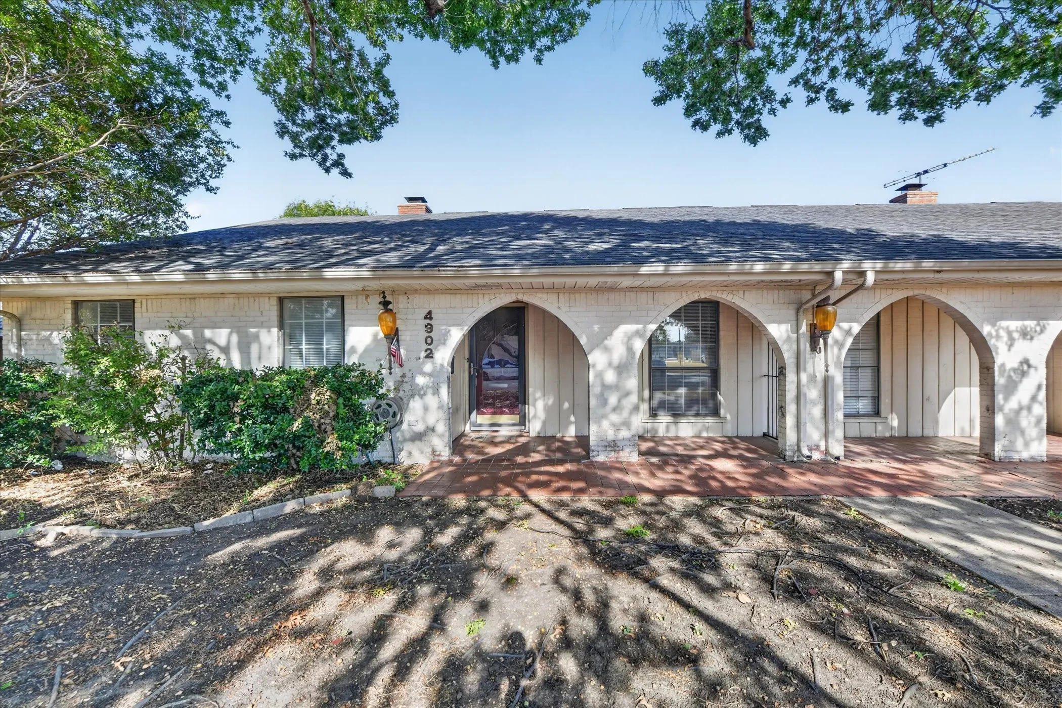 White brick exterior with arched entryway, dark shingles, and a red brick patio