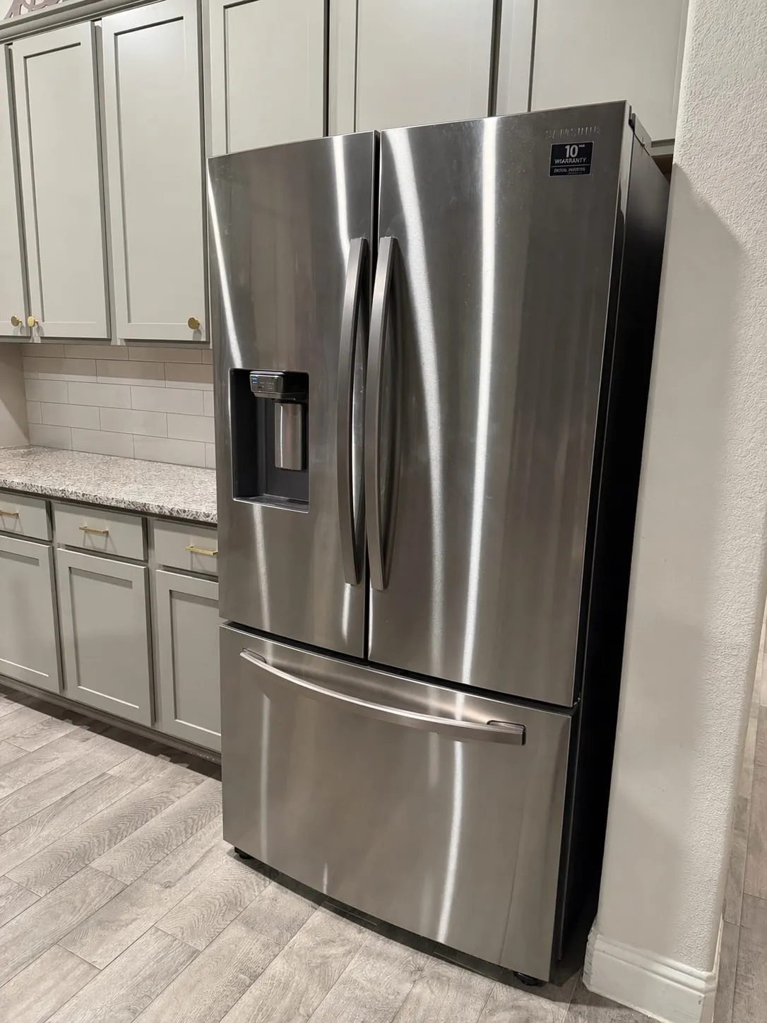 Stainless steel French door refrigerator with an external ice and water dispenser, complemented by light gray cabinetry, a speckled countertop, and a white subway tile backsplash
