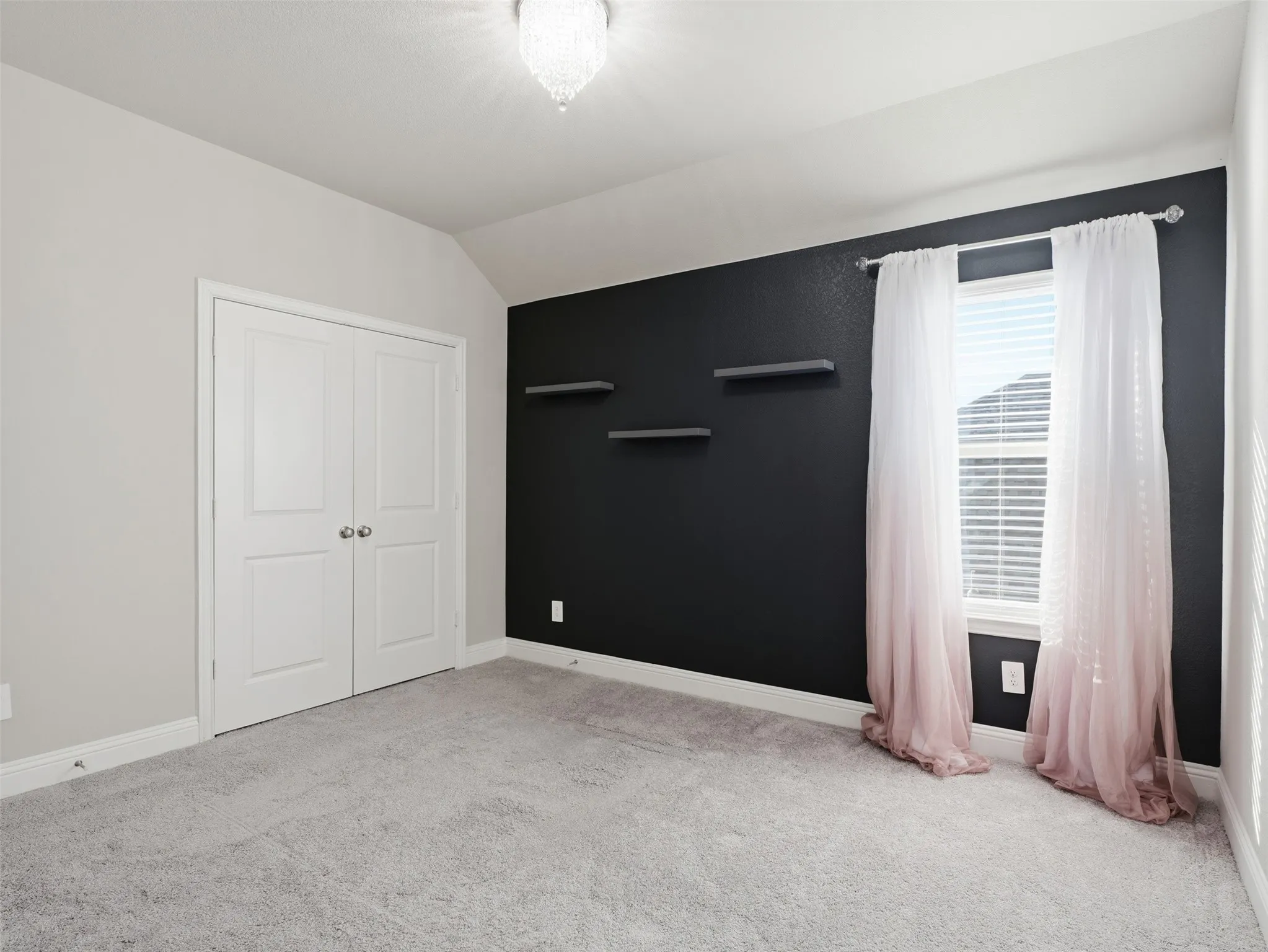 Spacious bedroom featuring neutral carpeting, a dramatic black accent wall with floating shelves, white double-door closet, and large window with pink curtains and plantation shutters
