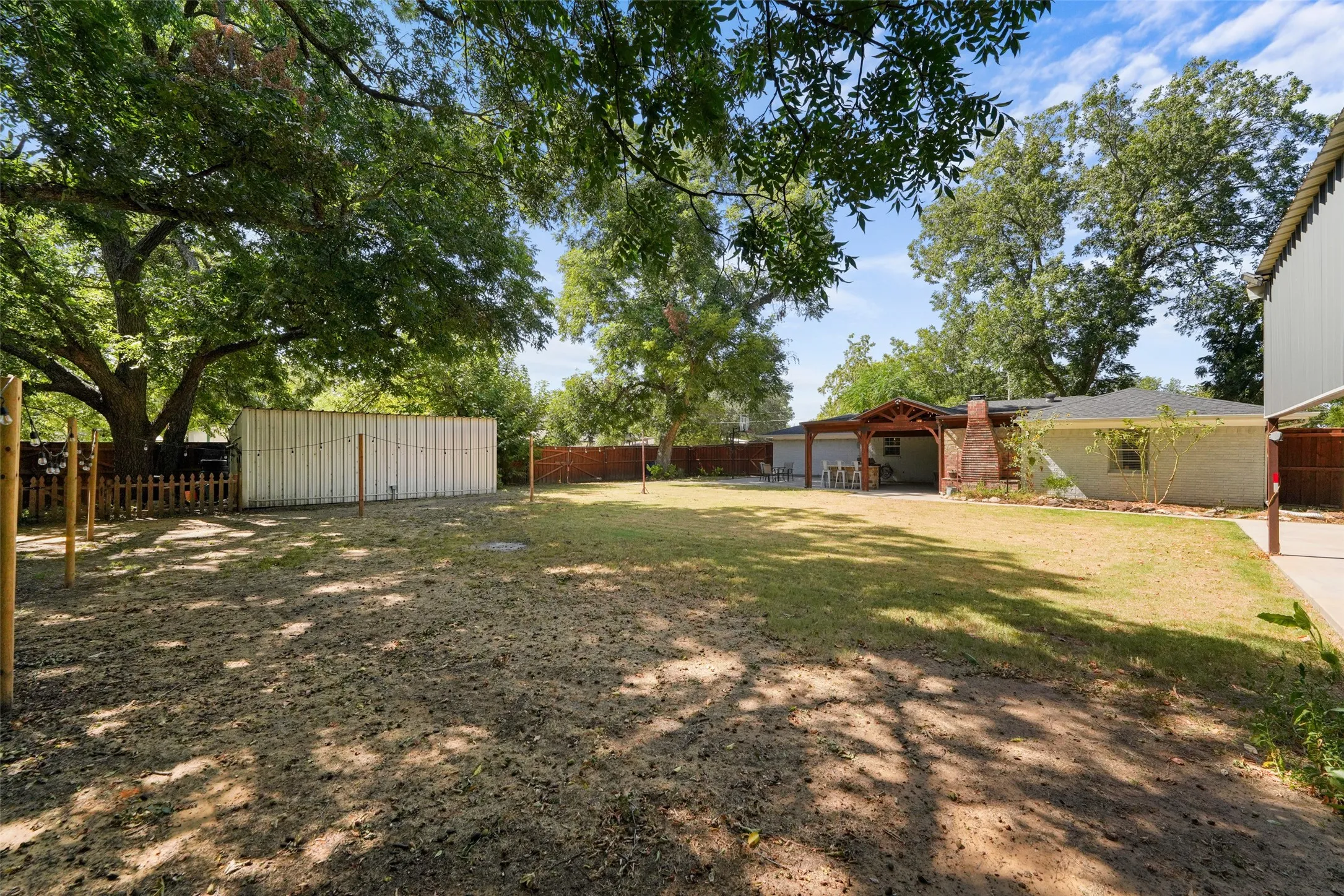 Expansive backyard featuring a covered patio with exposed wood beams, a brick chimney, mature trees, and a storage structure