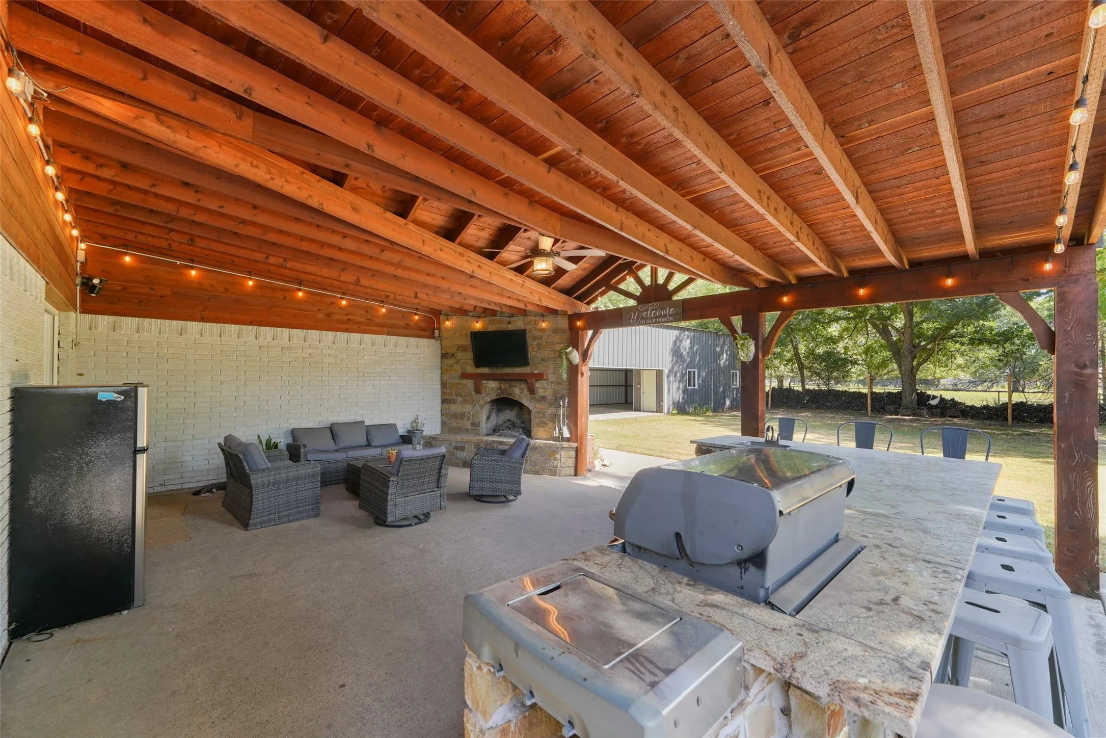 Expansive covered patio featuring a built-in outdoor kitchen with grill, wood-beamed ceiling, stone fireplace, and concrete flooring