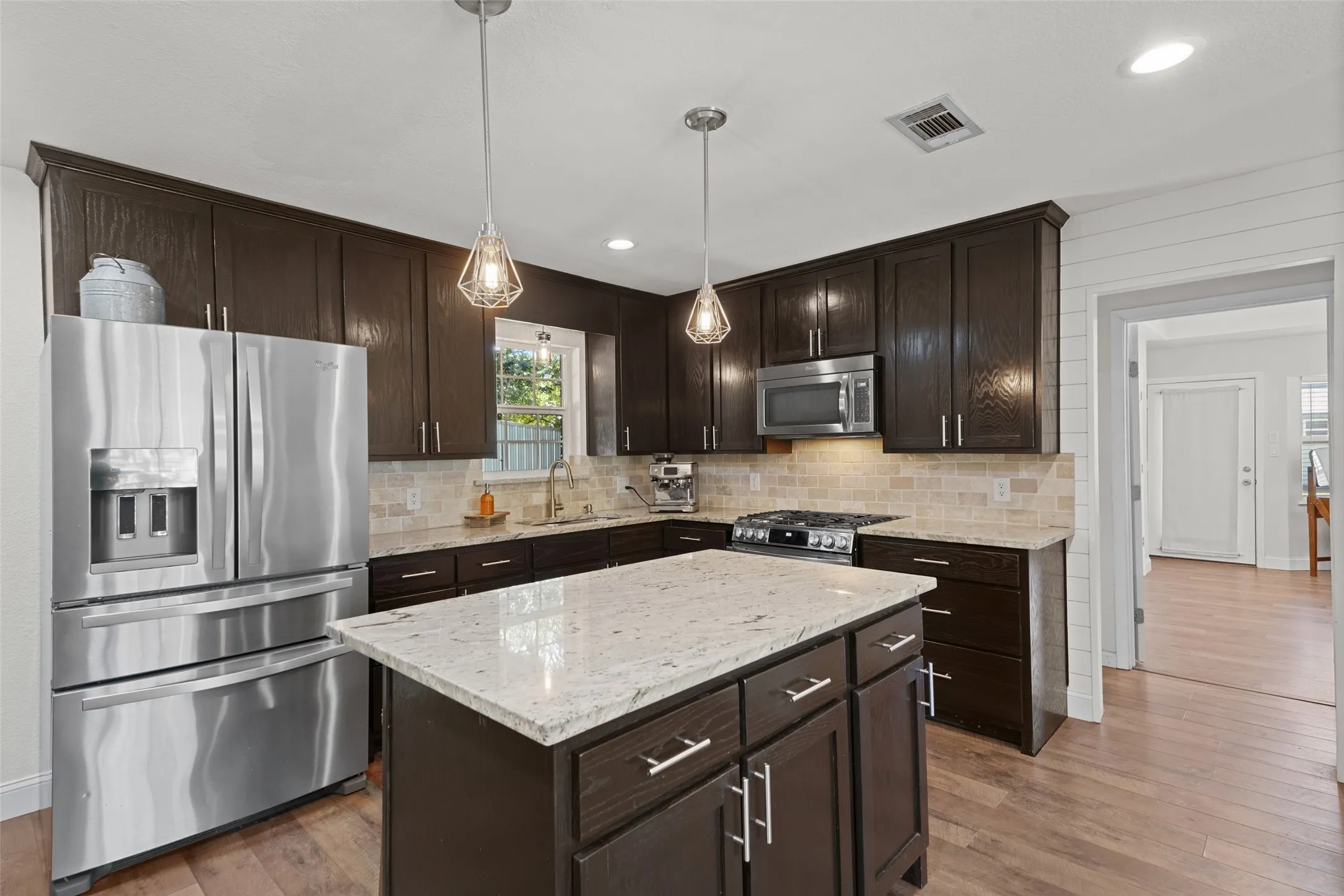 Kitchen featuring dark wood cabinetry, a center island with a light-toned countertop, stainless steel appliances, a tile backsplash, and wood-finish flooring