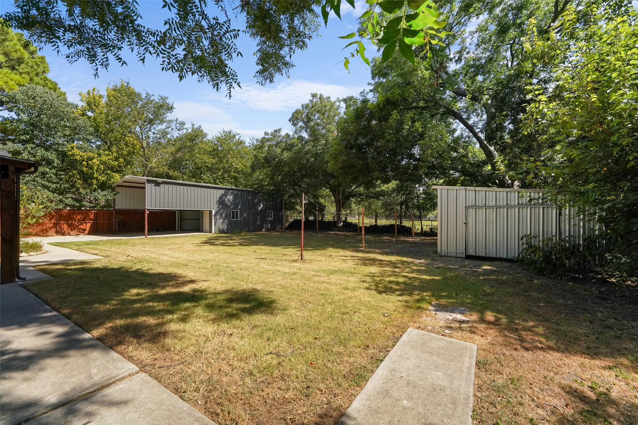Expansive yard space featuring a detached metal structure, a separate storage shed, concrete pathways, and mature trees