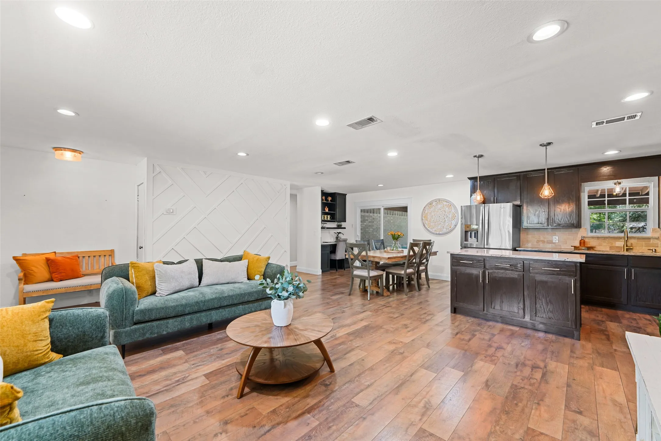 Open-concept living area featuring wood-finish flooring, recessed lighting, a decorative accent wall, a kitchen island with pendant lighting, and stainless steel appliances