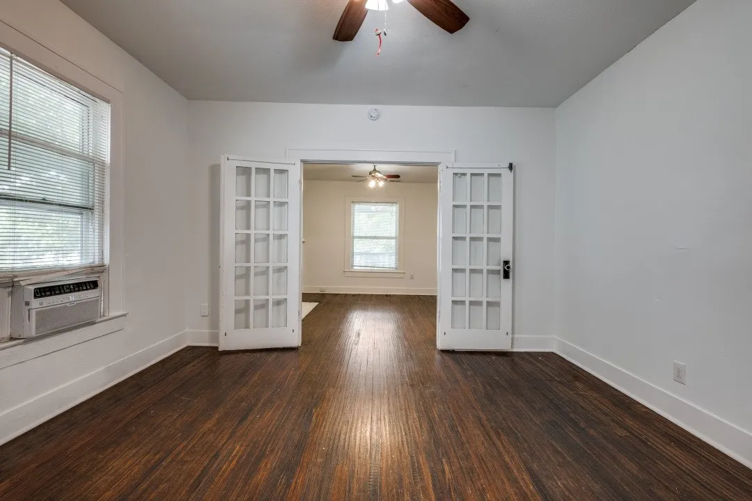 Hardwood flooring, white walls, double French doors, large window with blinds, and a ceiling fan
