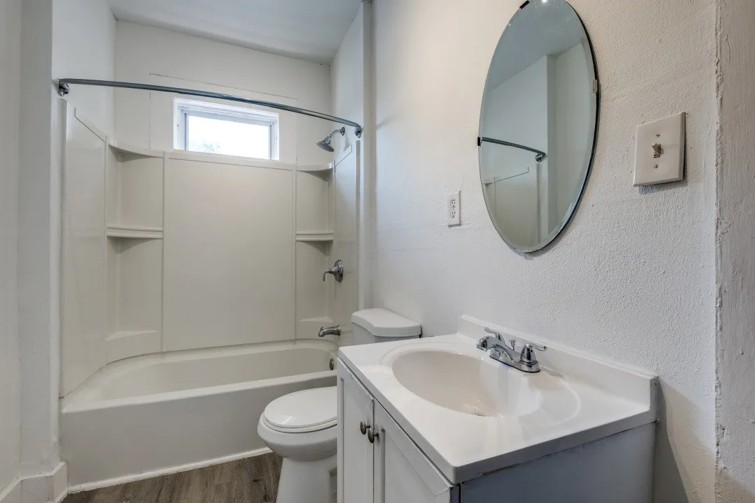 Bathroom featuring a white vanity with integrated sink, chrome fixtures, oval wall mirror, full-size bathtub with shower, and wood-finish flooring