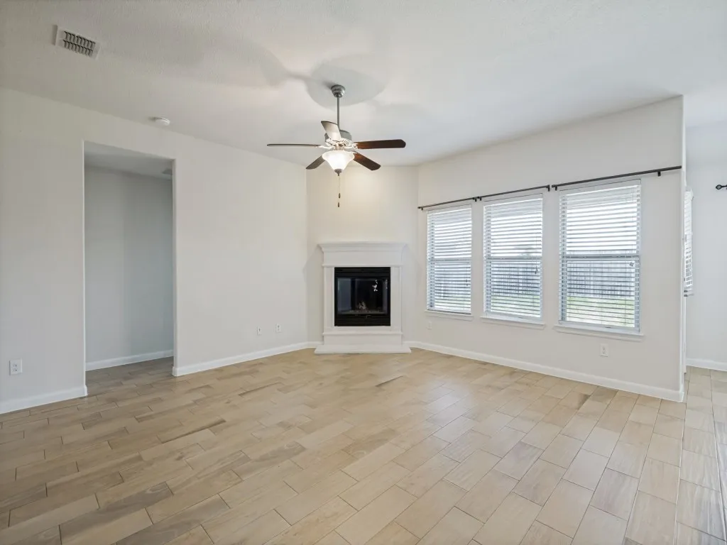 Wood-finish tile flooring throughout, featuring a corner fireplace with a white mantel, ceiling fan with integrated lighting, and multiple windows with horizontal blinds