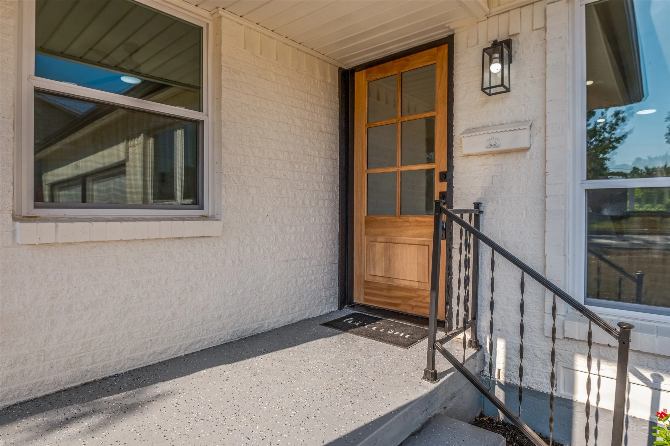 Wood-panel entry door with glass inserts, textured exterior wall finish, black wrought iron railing, contemporary outdoor sconce, and a covered porch