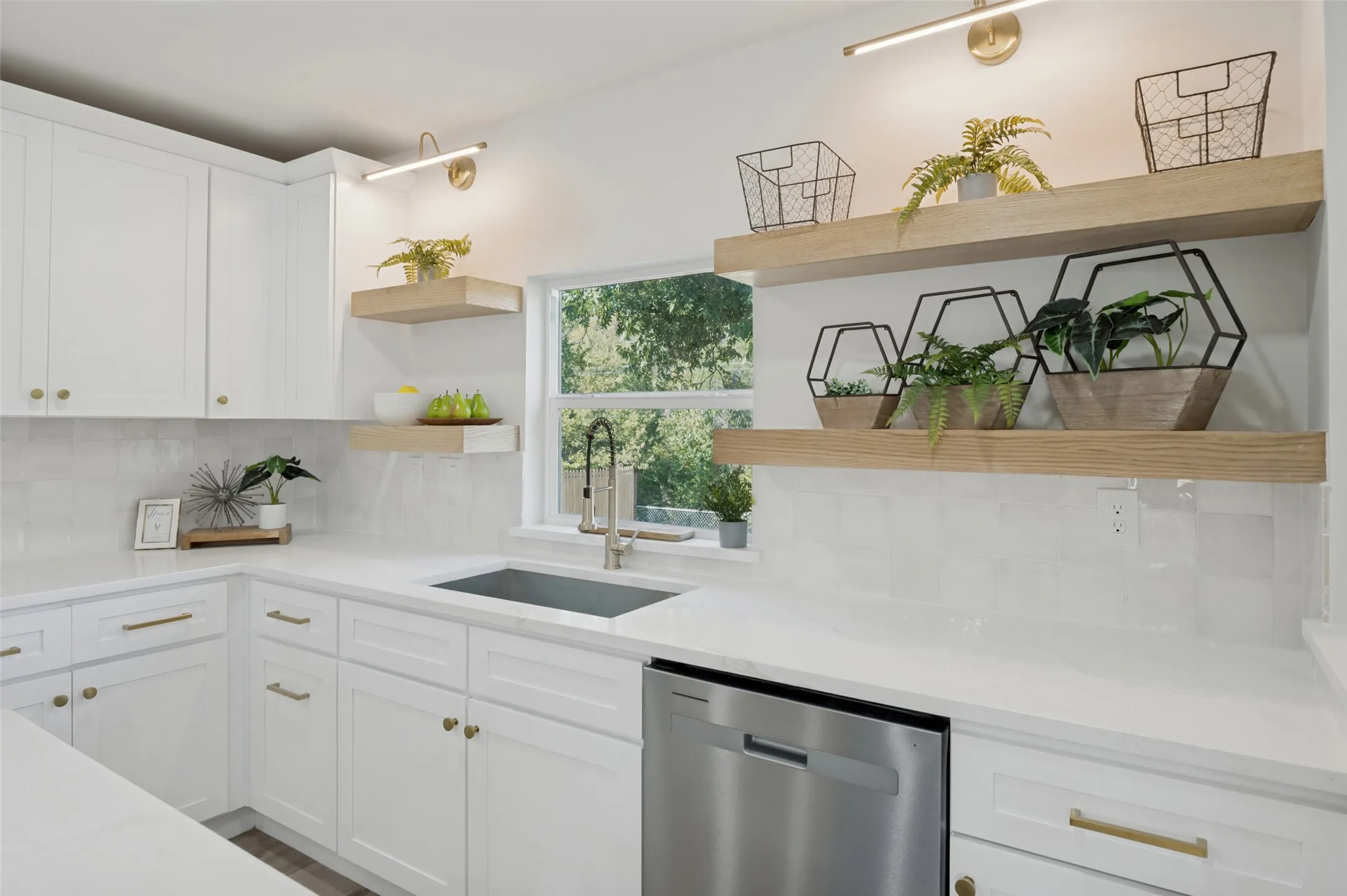White shaker-style cabinetry with gold hardware, light-toned countertops, white subway tile backsplash, stainless steel dishwasher, and open wood-finish shelving