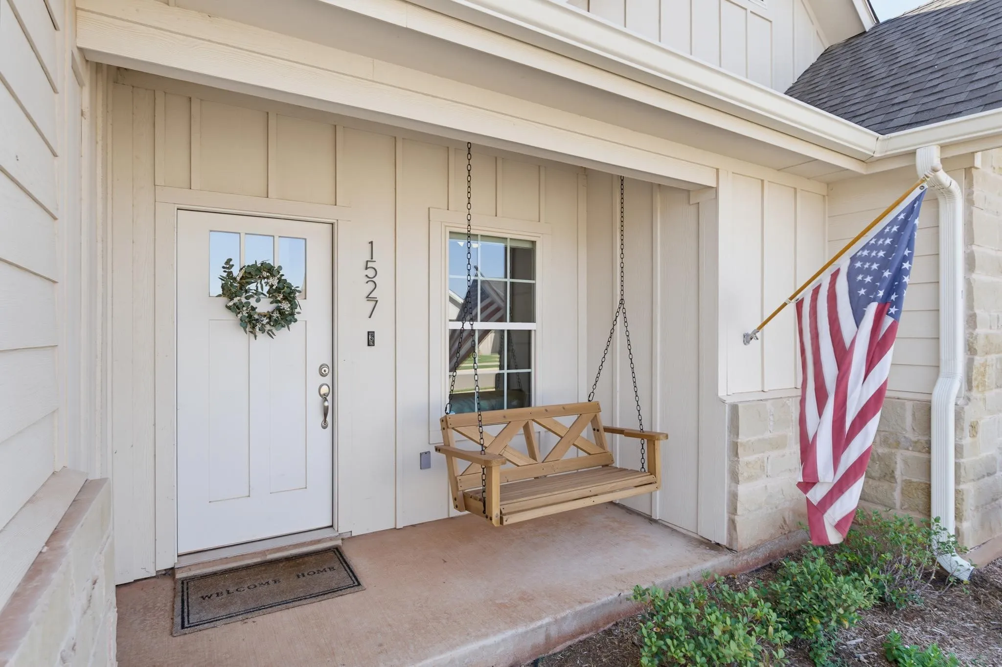 Covered concrete front porch featuring a wooden porch swing, board and batten siding, stone facade accents, and a white front door with windowpanes