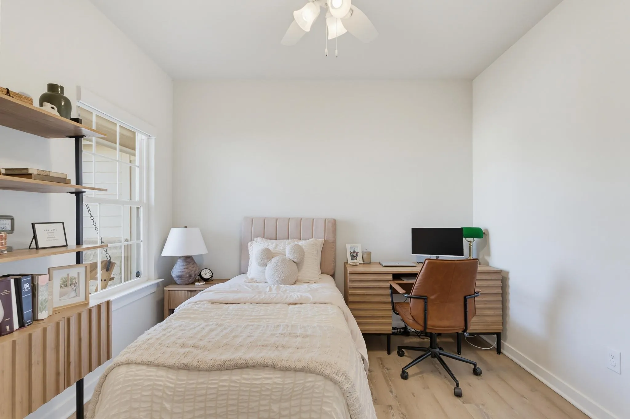 Bright bedroom featuring warm wood-finish flooring, a built-in shelving unit with open storage, and a dedicated workspace with desk and chair