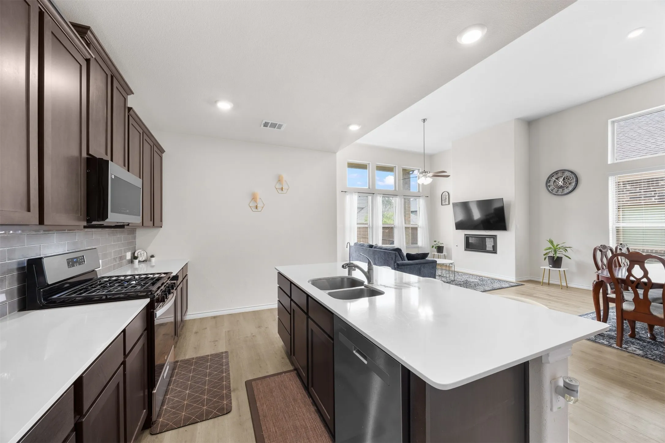 Open-concept design featuring a kitchen island with a double sink, dark wood-finish cabinetry, light wood-finish flooring, and a living area with a fireplace