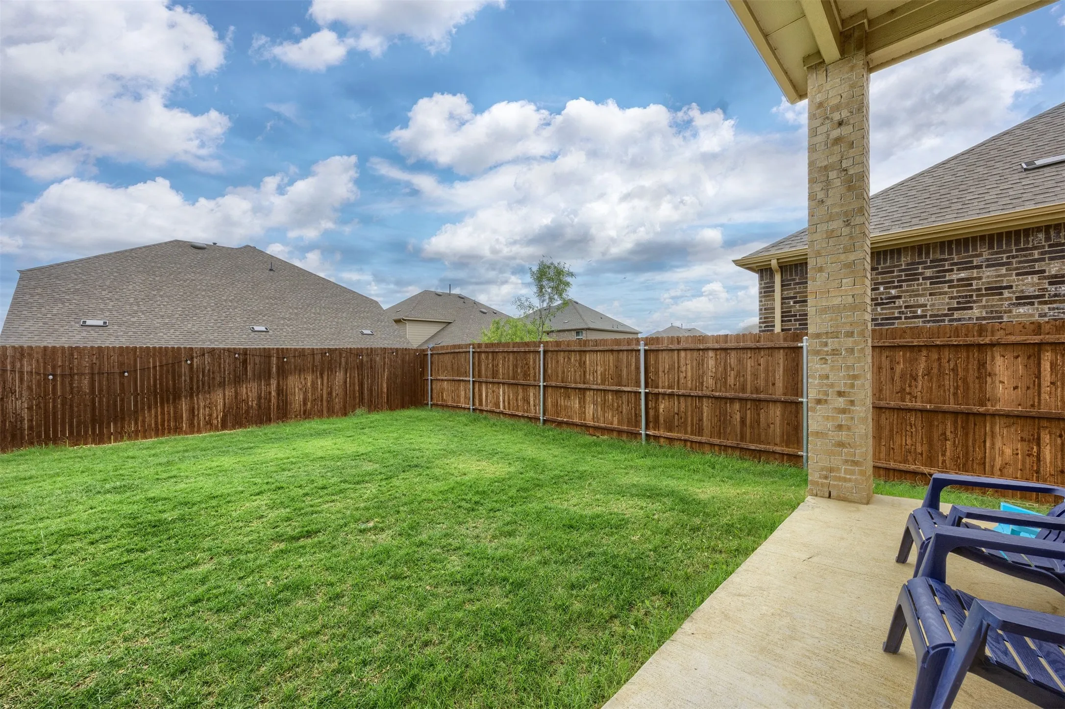 Expansive backyard featuring a manicured lawn, a stained wood privacy fence, and a covered concrete patio supported by a brick column