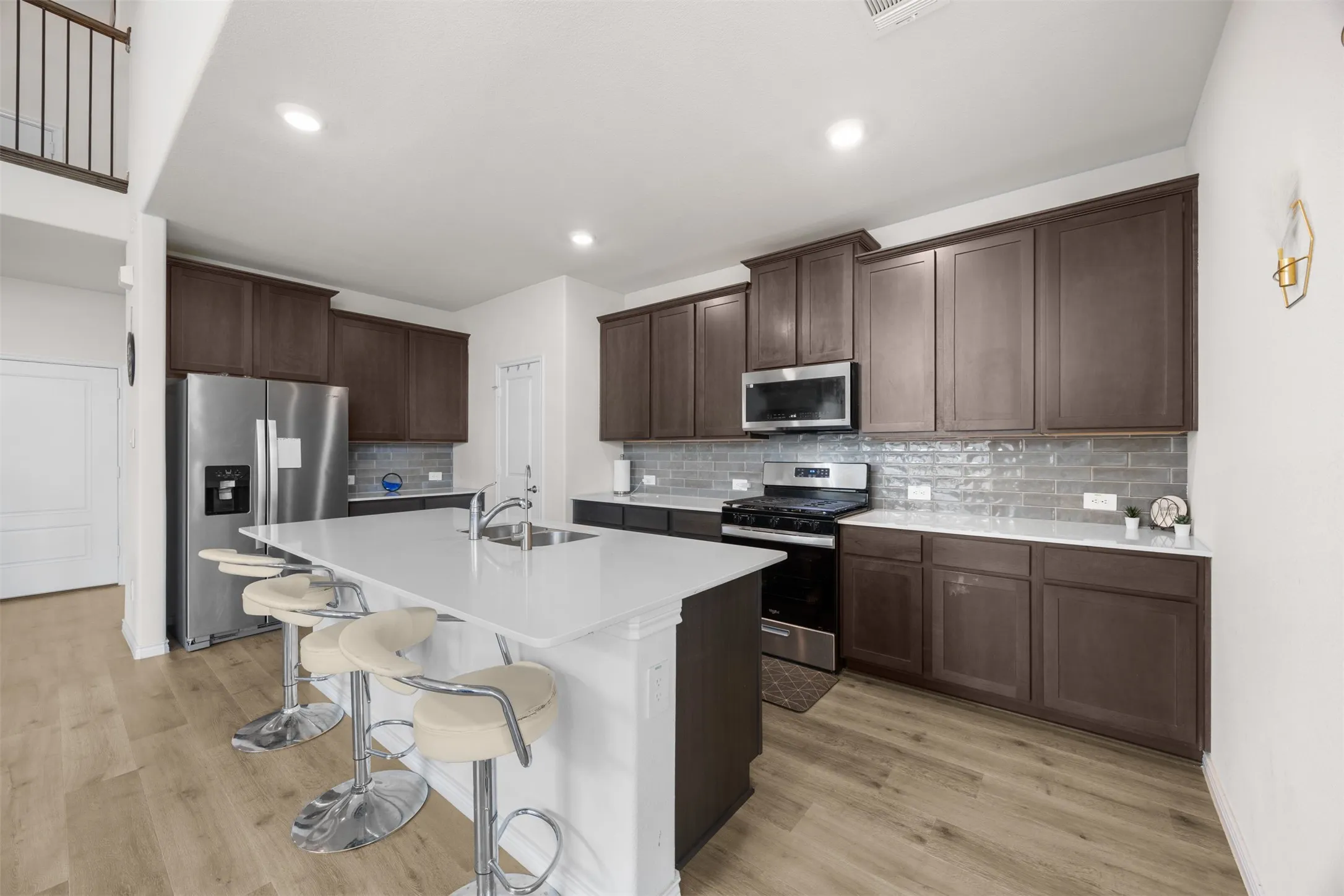 Kitchen featuring an expansive island with a built-in sink, wood-finish flooring, dark wood cabinetry, stainless steel appliances, and a tiled backsplash