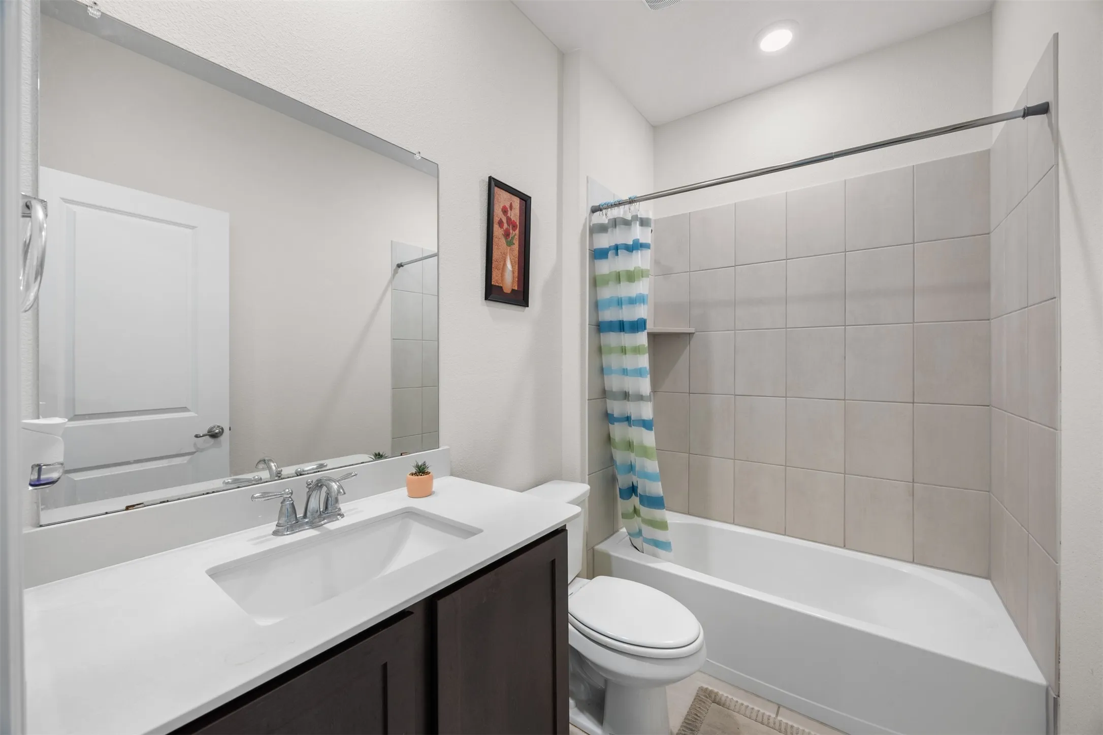 Bathroom featuring a built-in tub with neutral tile surround and recessed shelf, a single vanity with dark wood-finish cabinetry, a white countertop with integrated sink, and a large wall-mounted mirror