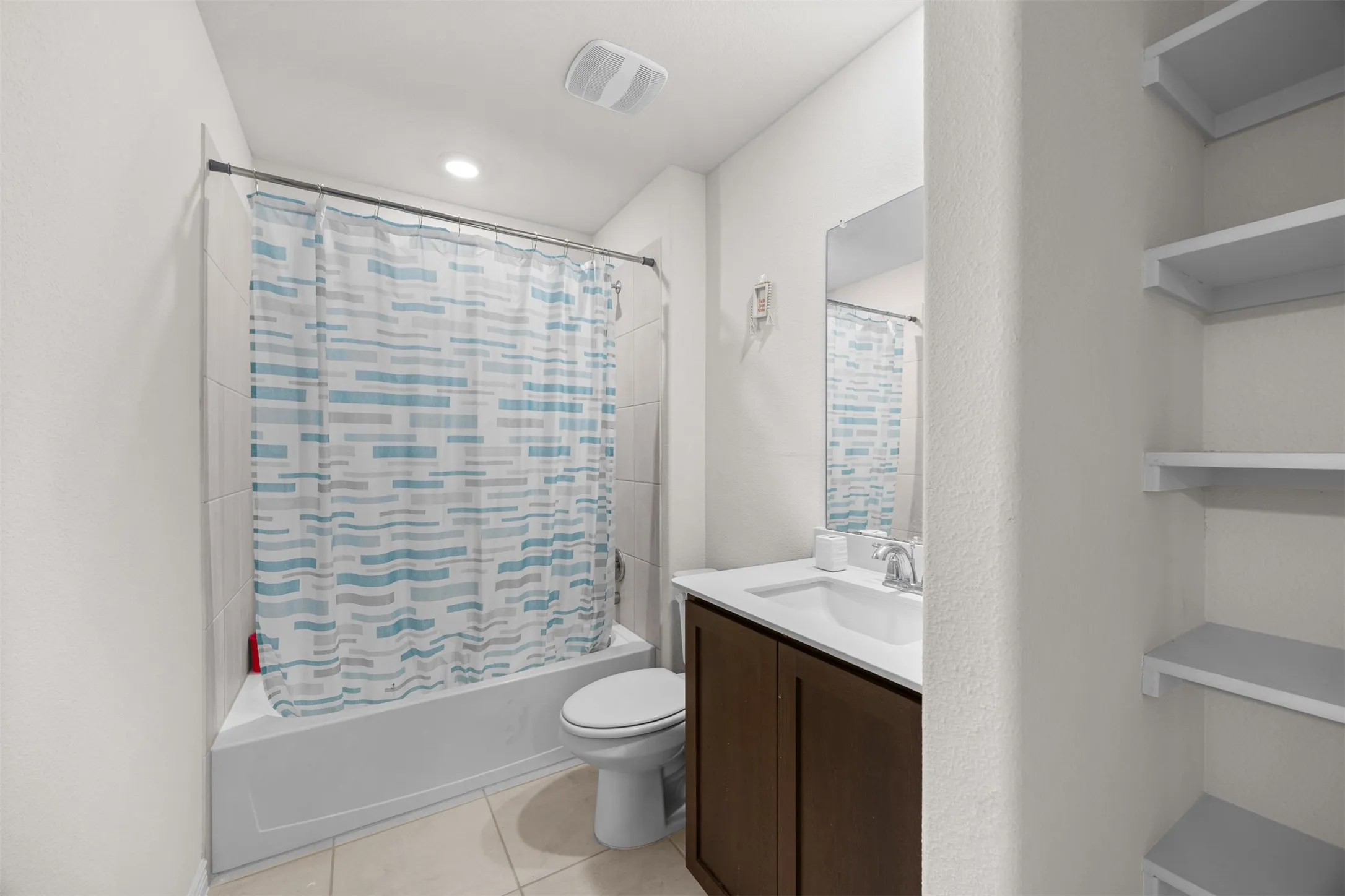 Full bathroom featuring a white tub, light-colored tile flooring, a dark wood-finish vanity with an integrated sink, and built-in shelving