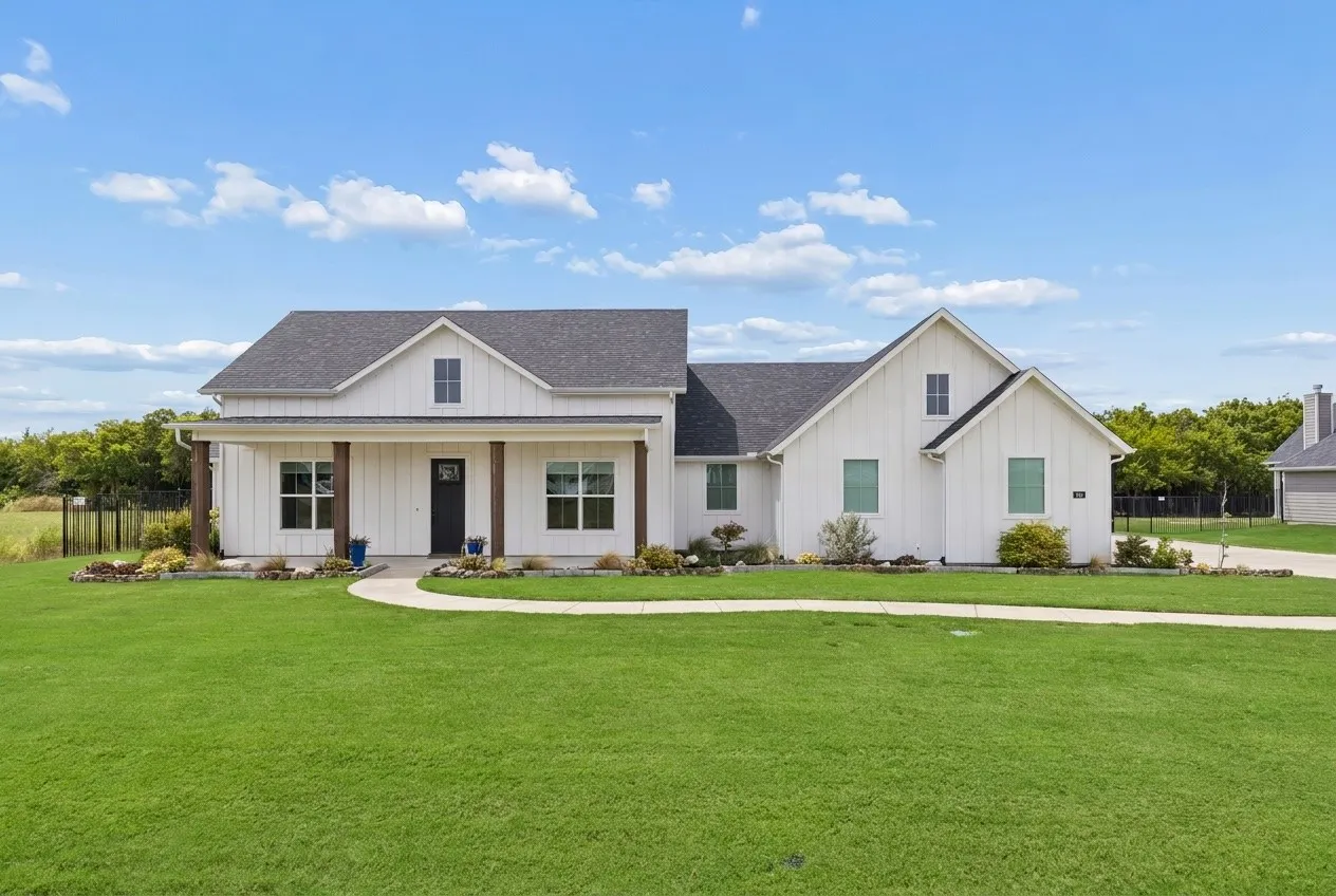 Modern farmhouse exterior with white board-and-batten siding, dark shingle roof, covered front porch with wood columns, and expansive lawn