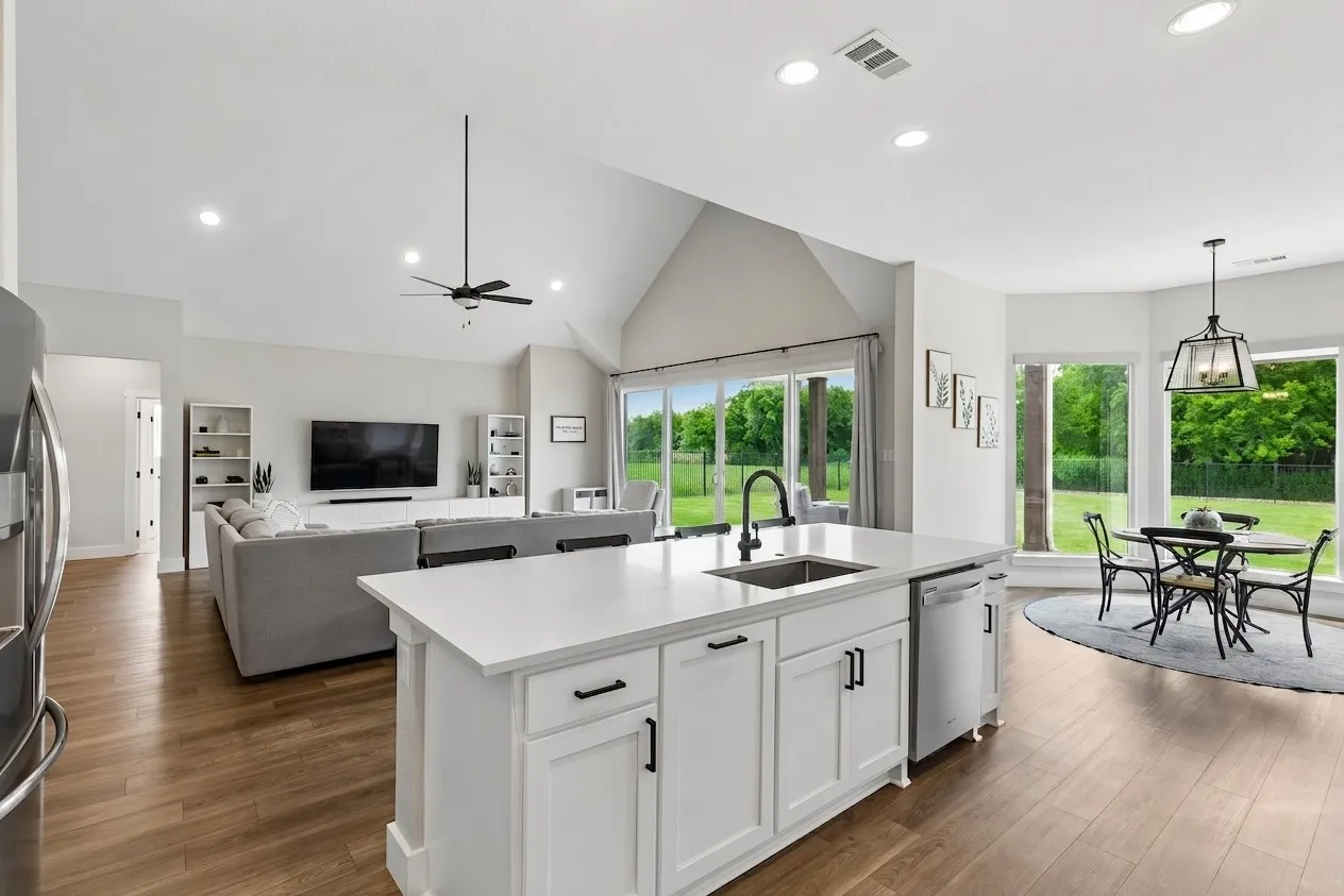 Open-concept living space featuring wood-finish LVP flooring, a kitchen island with a built-in sink, vaulted ceilings, and a dining area with large windows