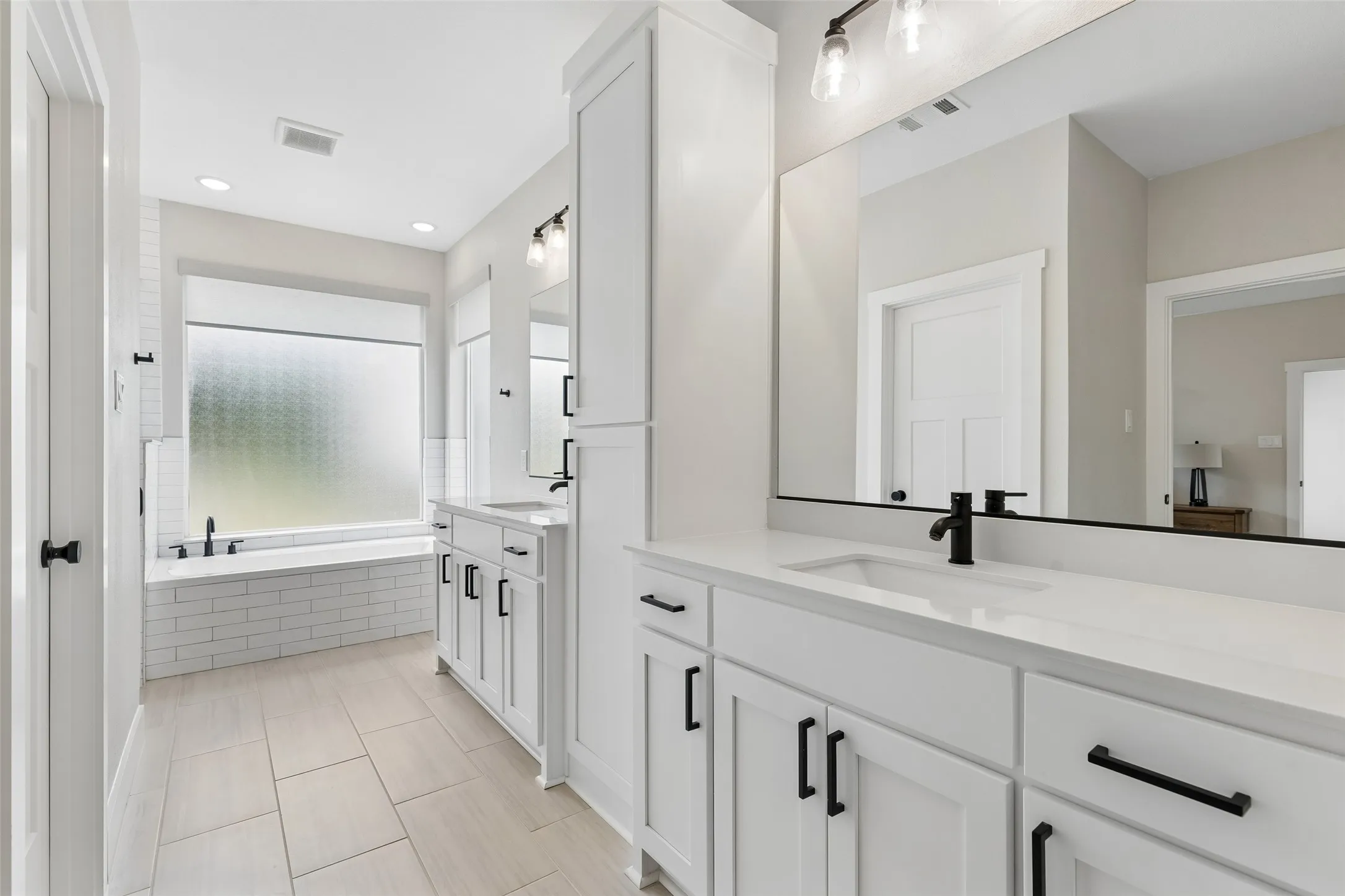 Dual vanities with white countertops, matte black fixtures, recessed lighting, a built-in tub with subway tile surround, and light-toned tile flooring