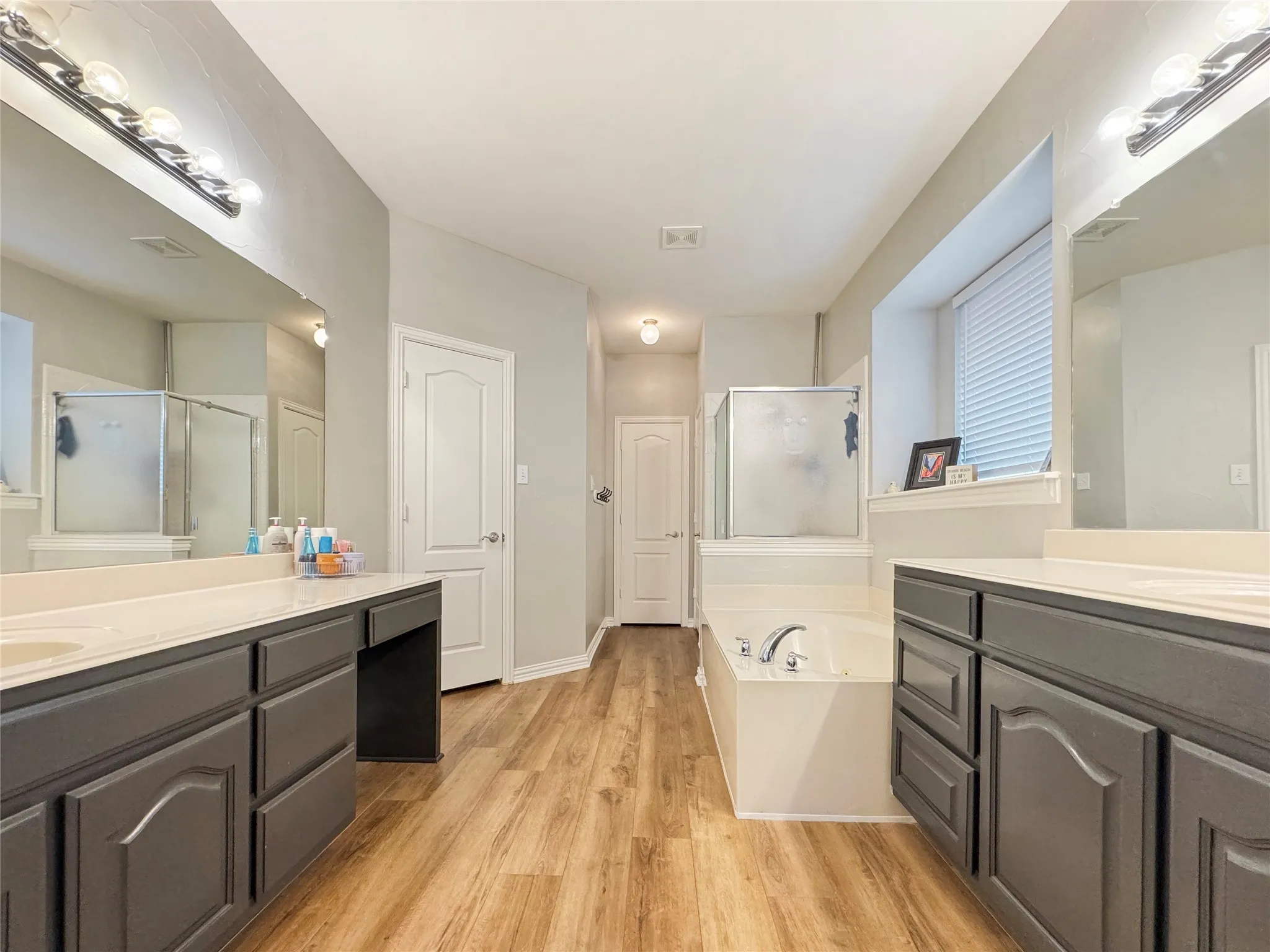 Dual vanities with white countertops, wood-finish flooring, a built-in jetted soaking tub, and a separate glass-enclosed shower