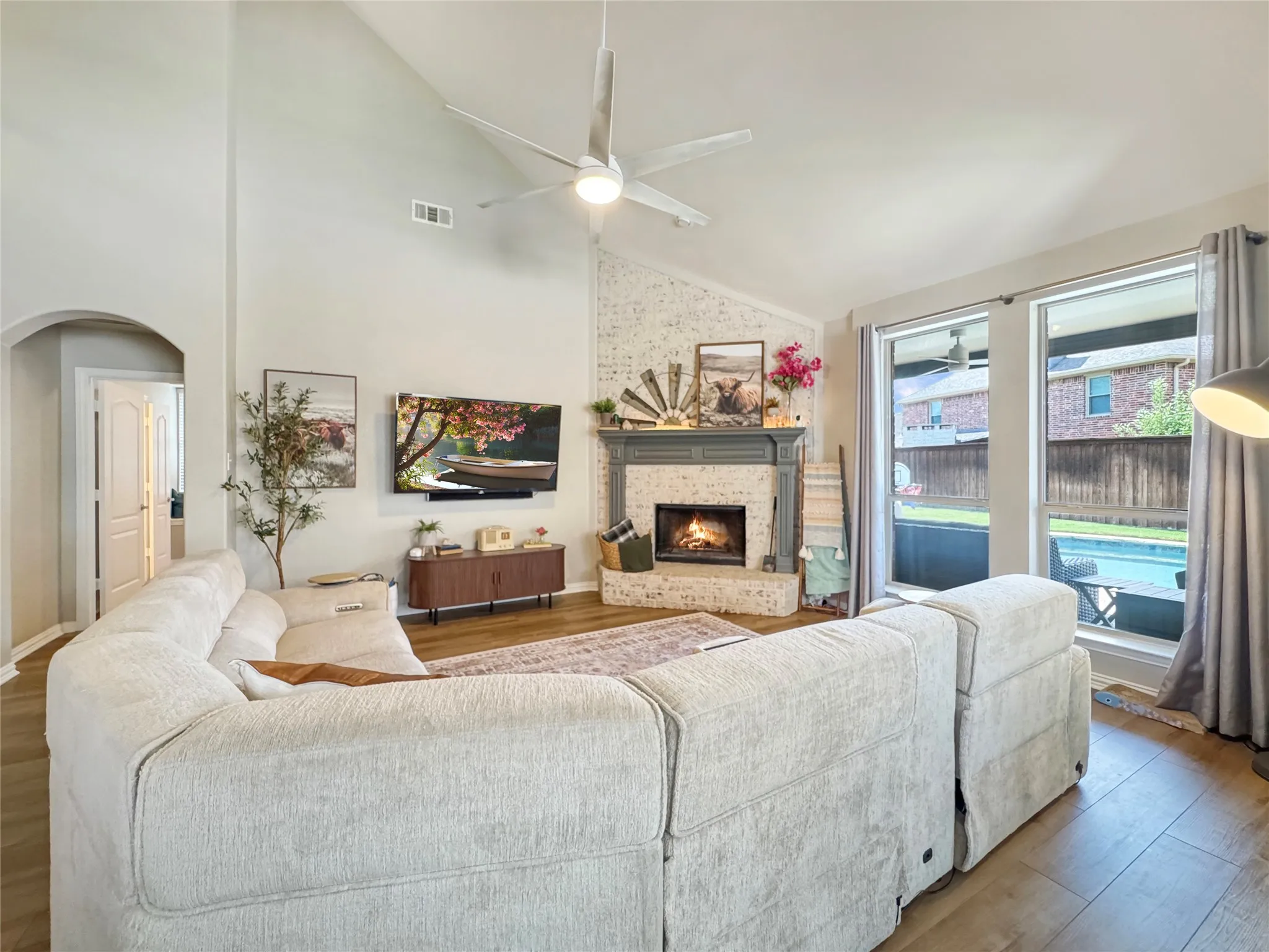 Living area featuring vaulted ceilings, wood-finish flooring, a brick fireplace with mantel, and large windows overlooking a backyard pool
