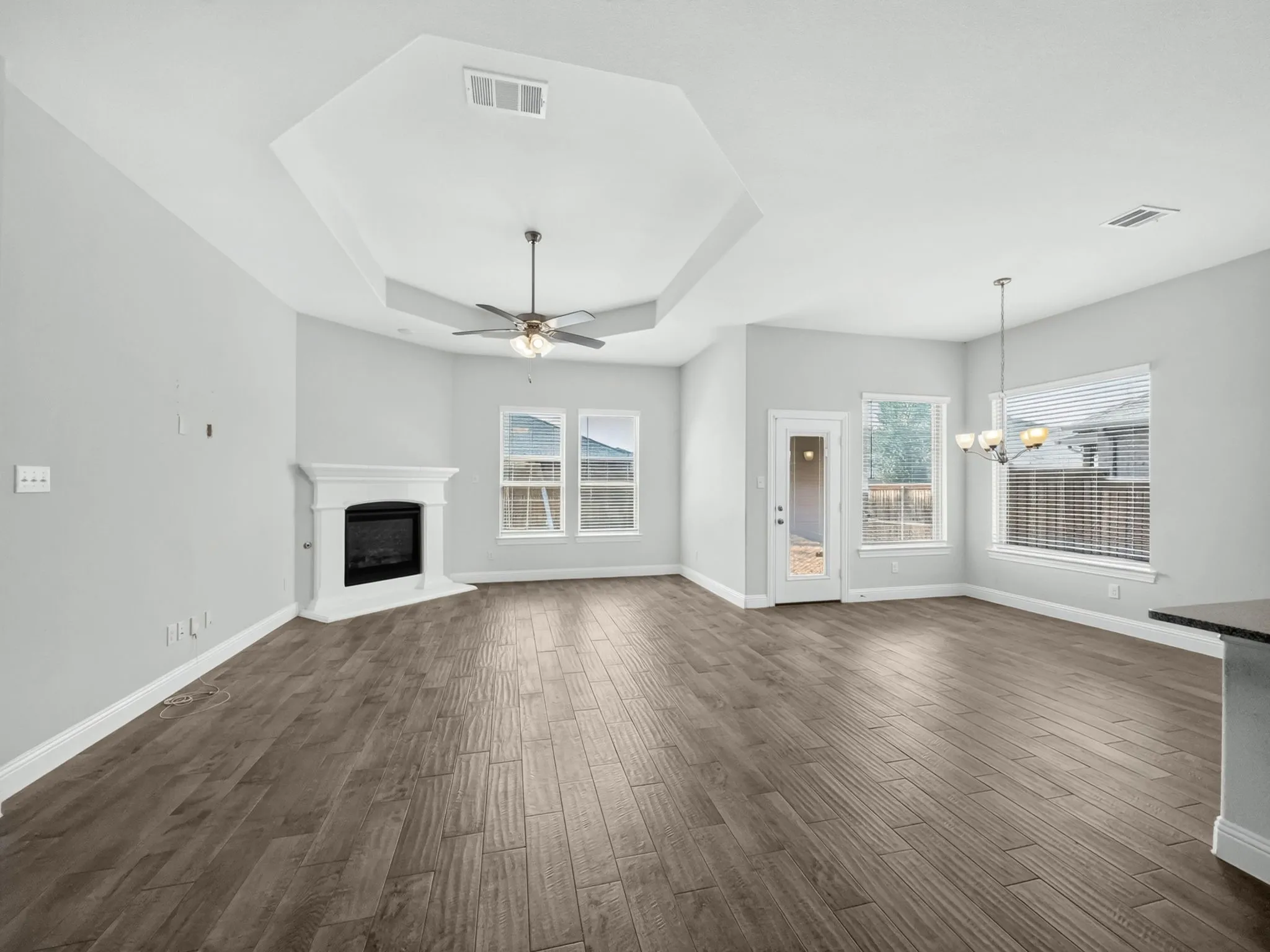 Wood-finish flooring throughout, a tray ceiling with a ceiling fan, a corner fireplace with a white mantel, multiple windows, and a glass-paneled exterior door