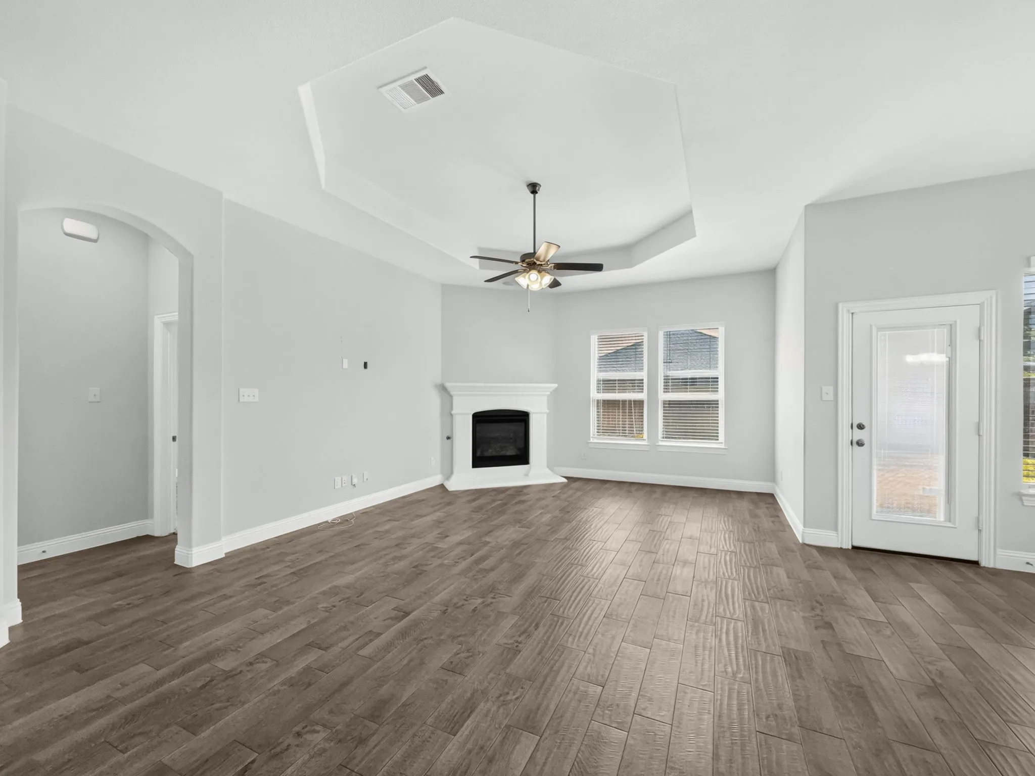Tray ceiling with a ceiling fan, wood-finish flooring, a white fireplace, multiple windows, and an arched entryway