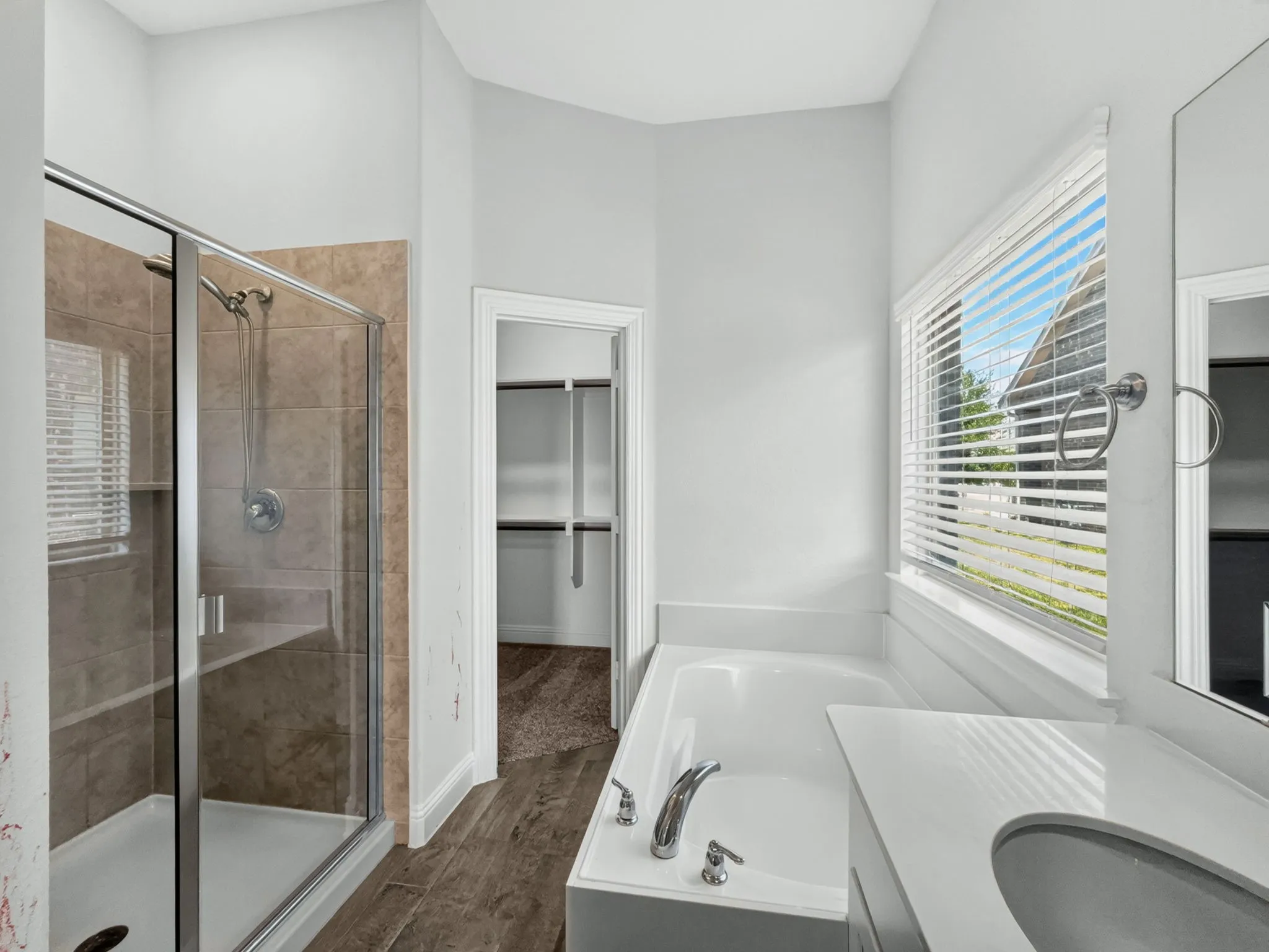 Bathroom featuring a glass-enclosed shower, soaking tub, window with blinds, and wood-finish flooring