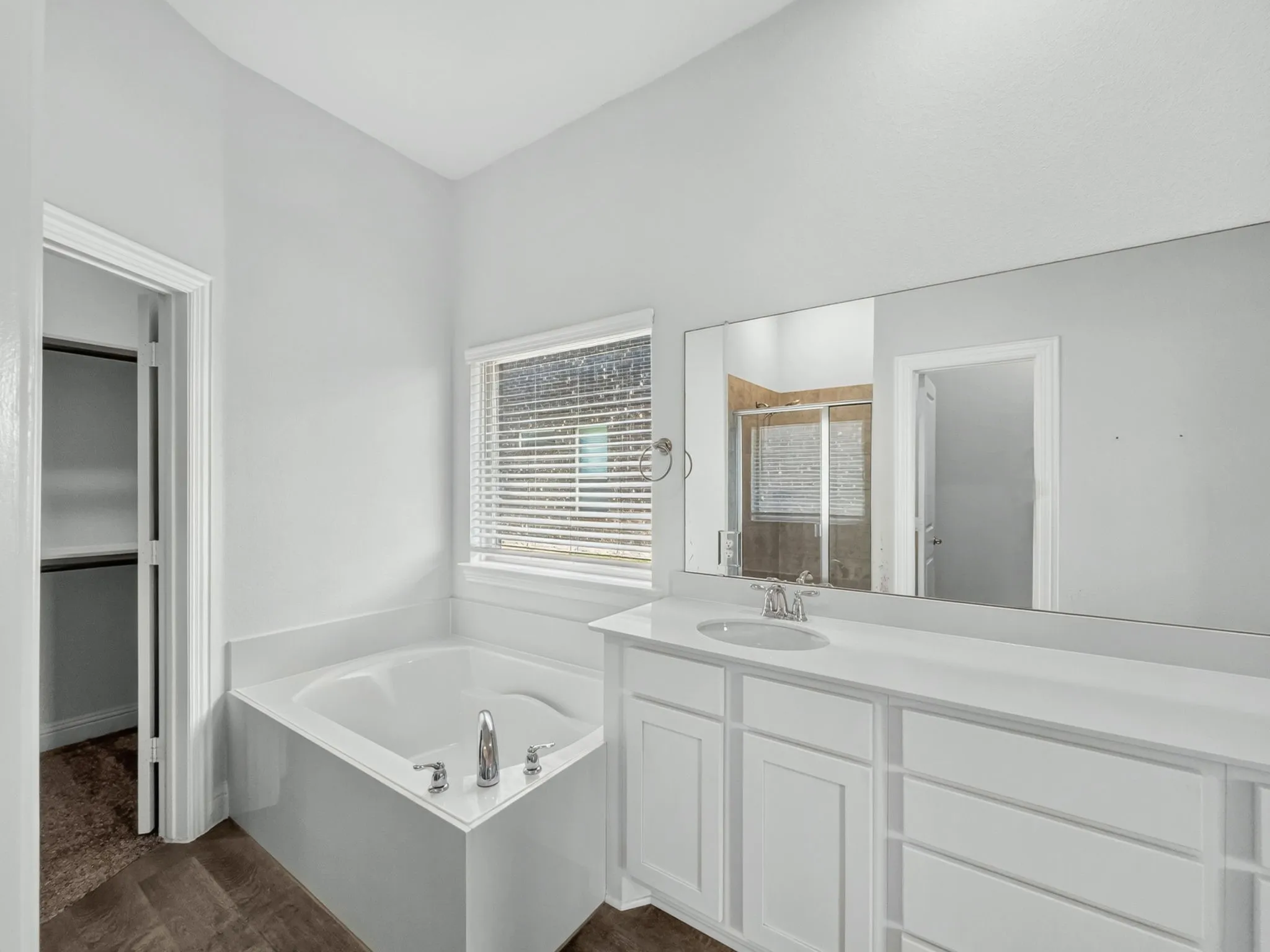 Corner soaking tub with deck-mounted chrome fixtures, white vanity with integrated sink, expansive vanity mirror, and wood-finish flooring