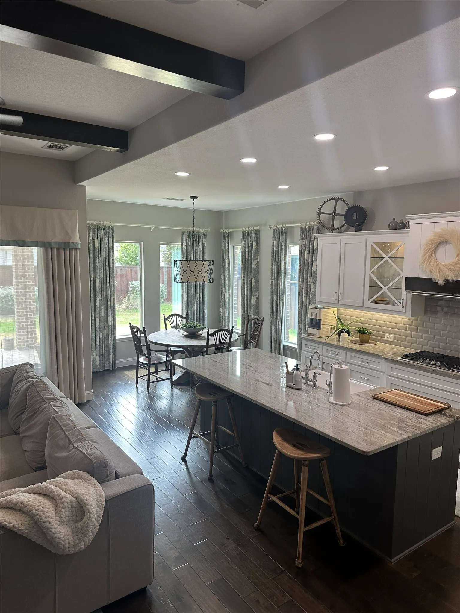 Spacious open-concept layout featuring dark wood-finish flooring, a kitchen island with stone countertop, recessed lighting, and decorative ceiling beams