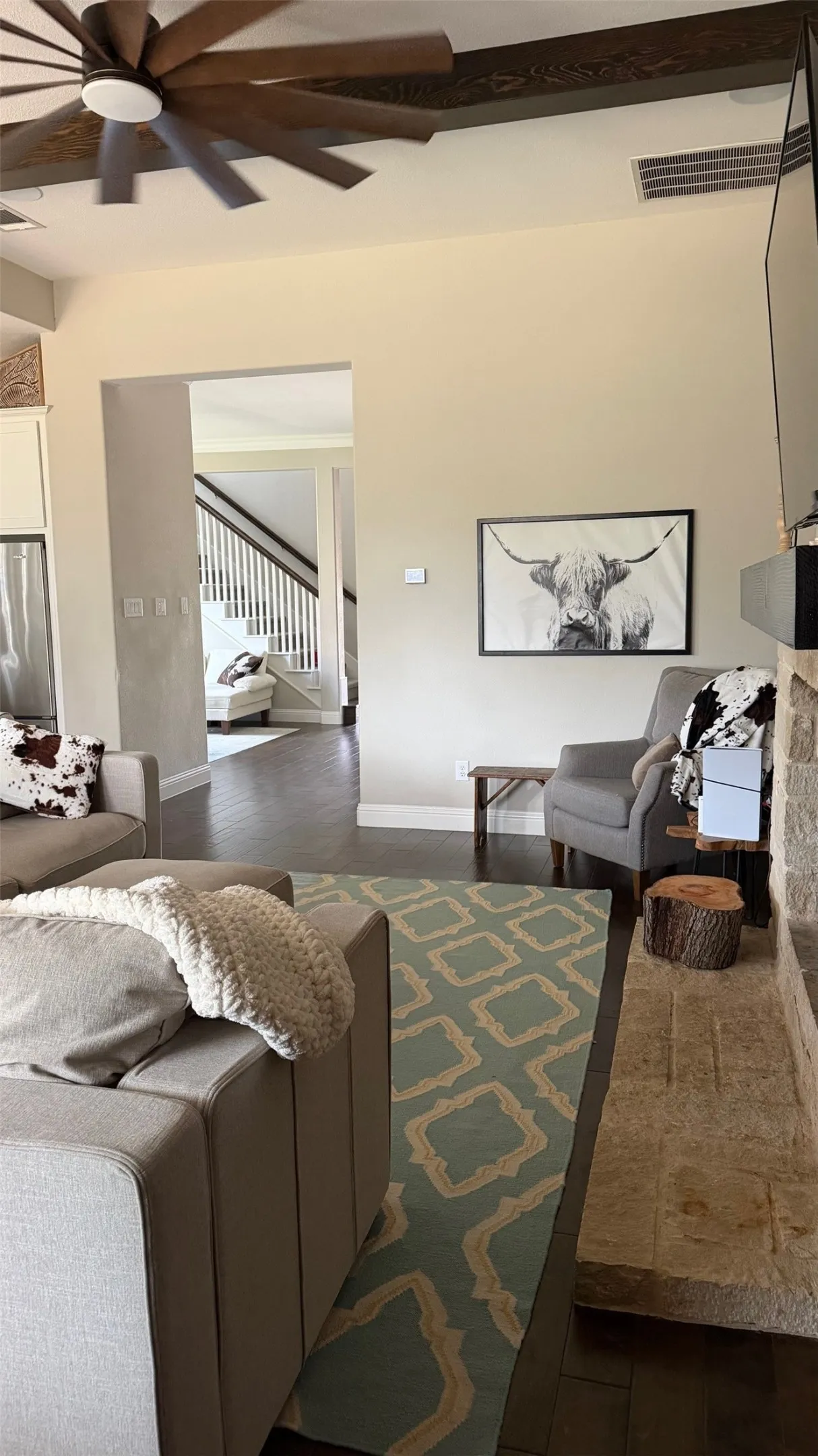 Dark wood-finish flooring with a stone fireplace, exposed ceiling beams, and a ceiling fan with integrated lighting