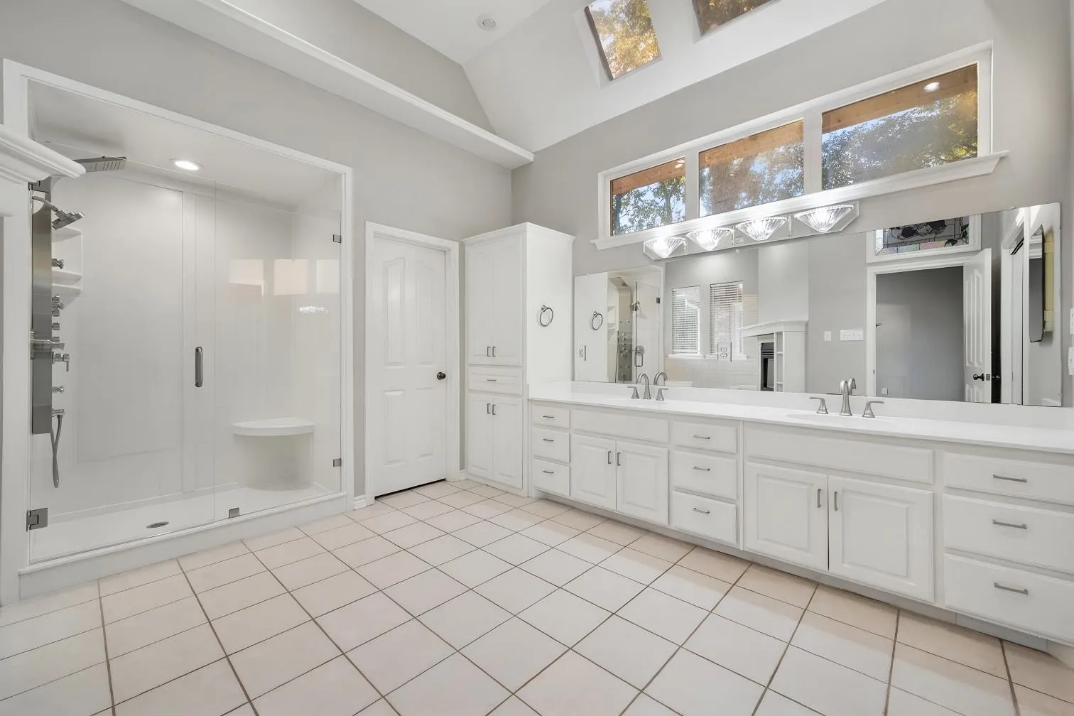 Dual vanity with white cabinetry and integrated sinks, glass-enclosed shower with built-in bench, vaulted ceiling, and transom windows