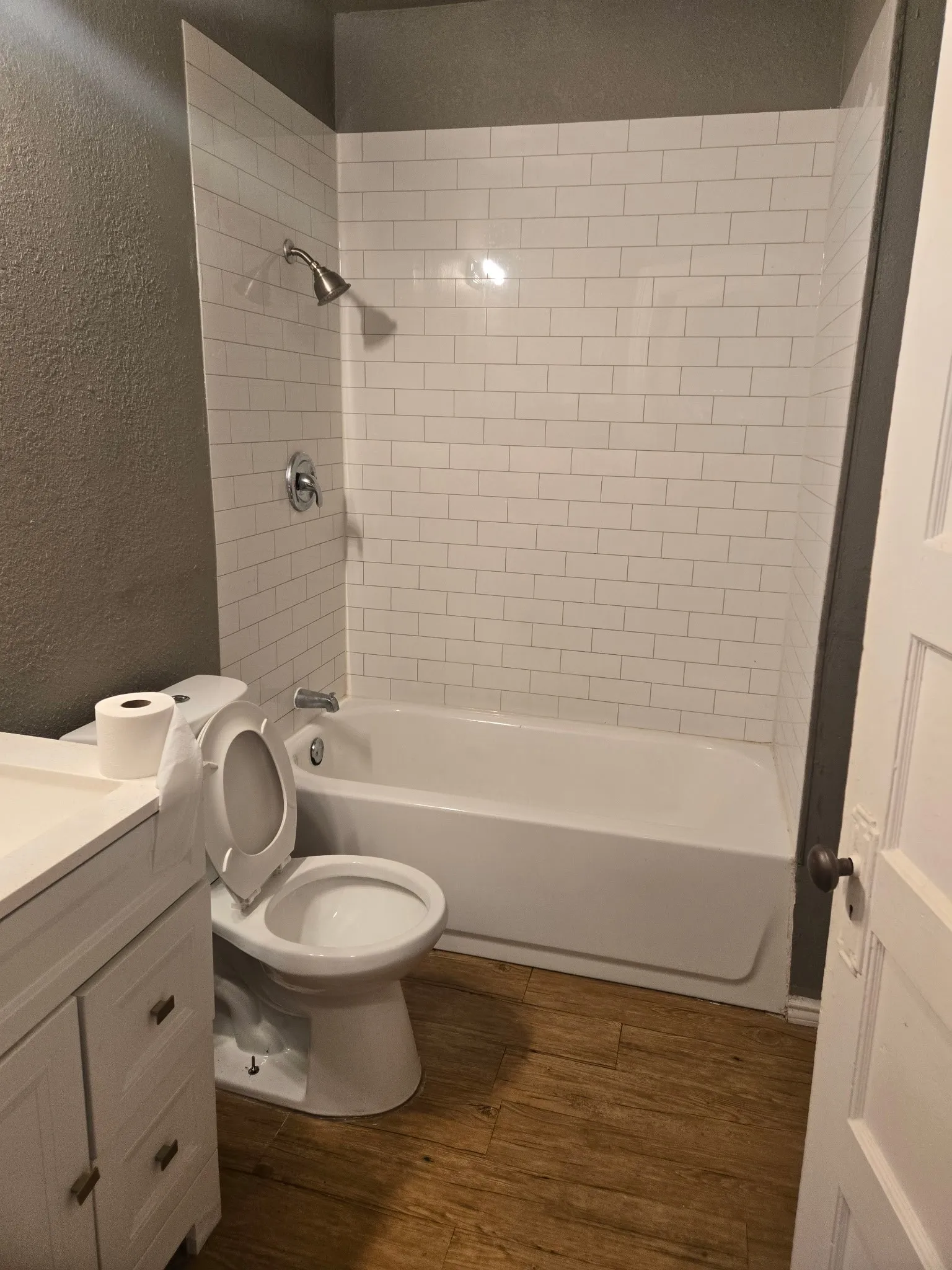 Bathroom featuring a white subway tile shower surround, a white bathtub, a white vanity with cabinet storage, a white toilet, and wood-finish flooring