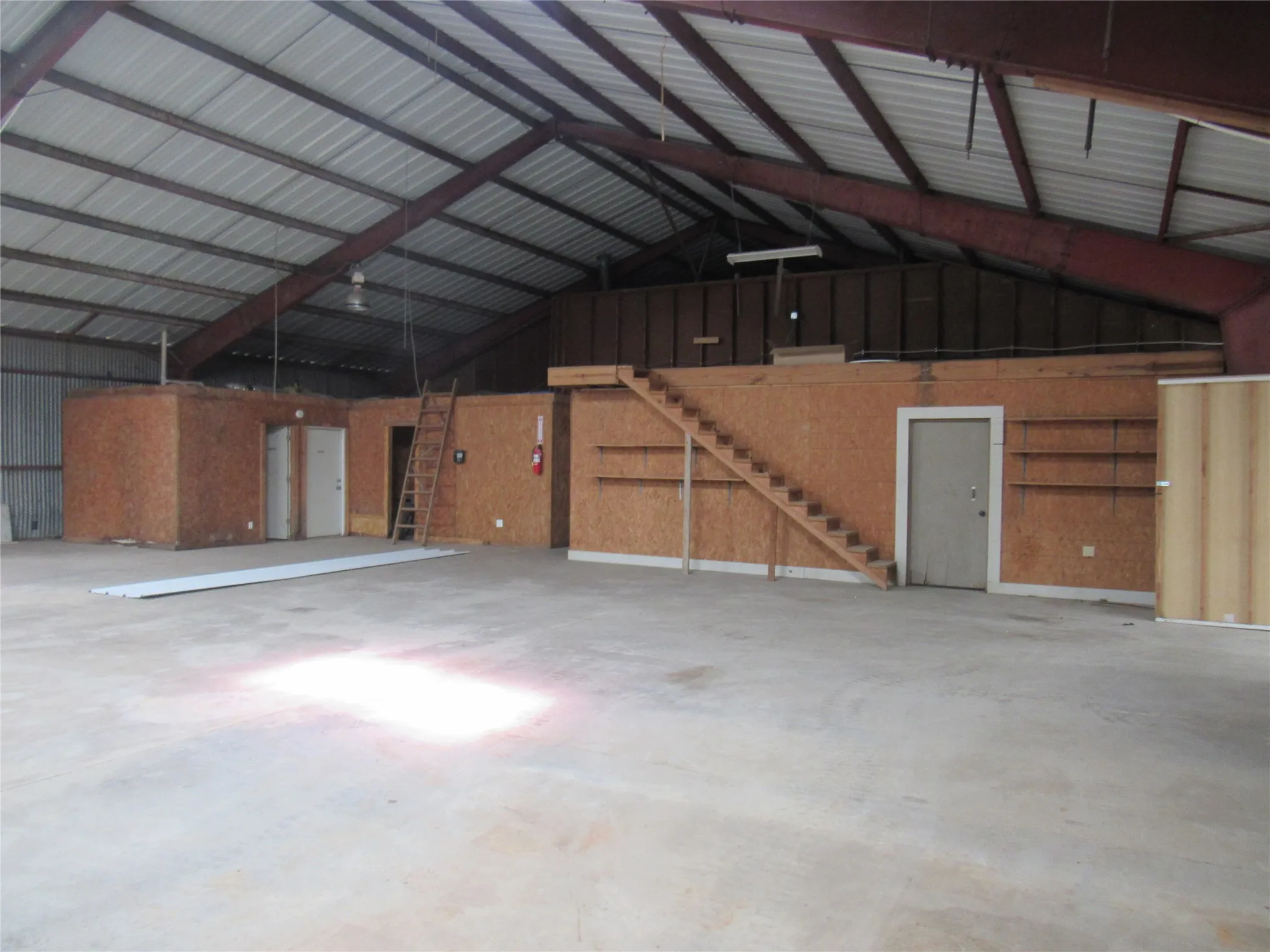 Expansive open interior space featuring a high-pitched metal roof, exposed steel beams, concrete flooring, built-in wooden shelving, and interior access doors