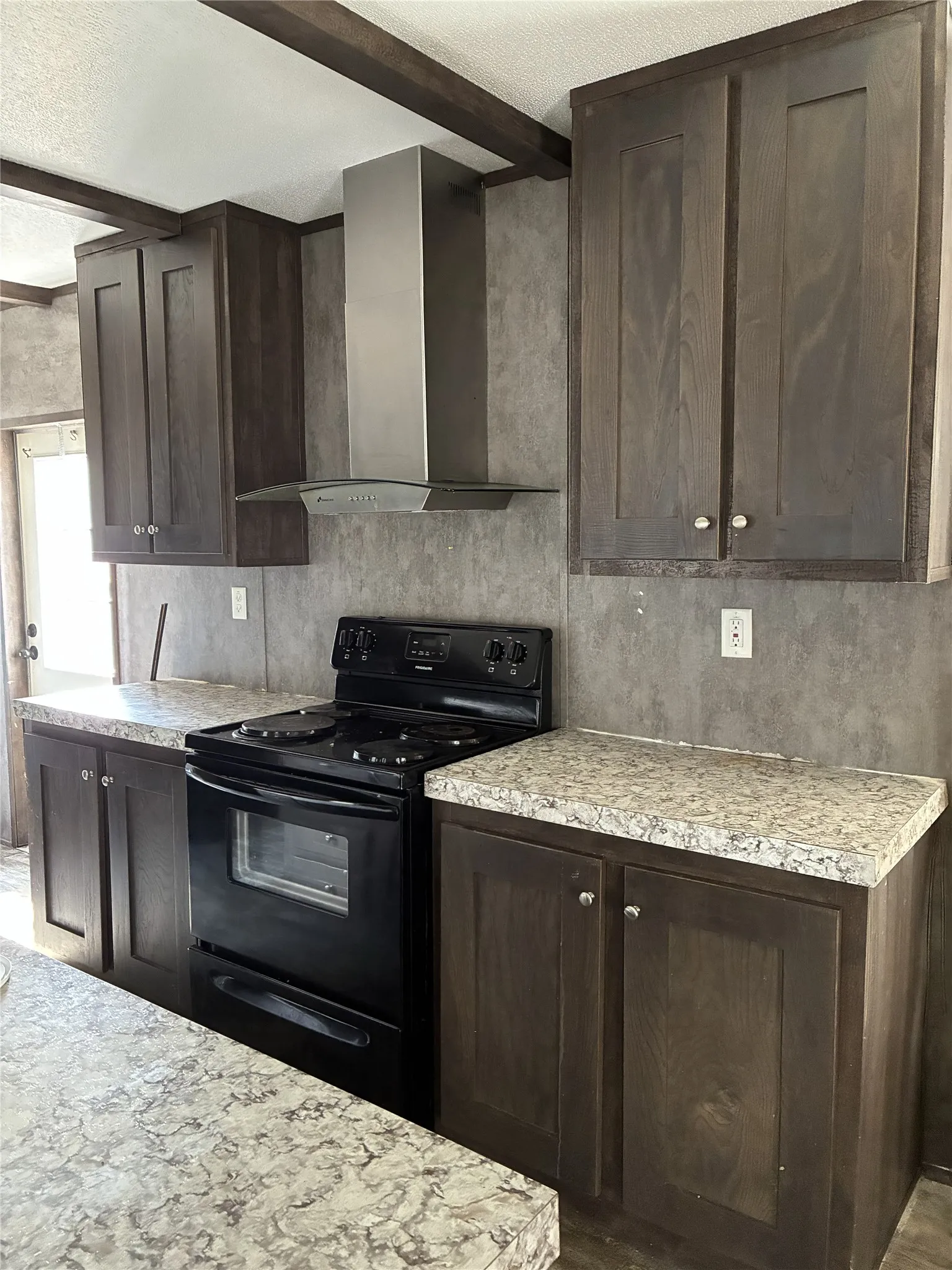 Dark wood-finish cabinetry with silver hardware, speckled countertops, stainless steel range hood, and black electric range