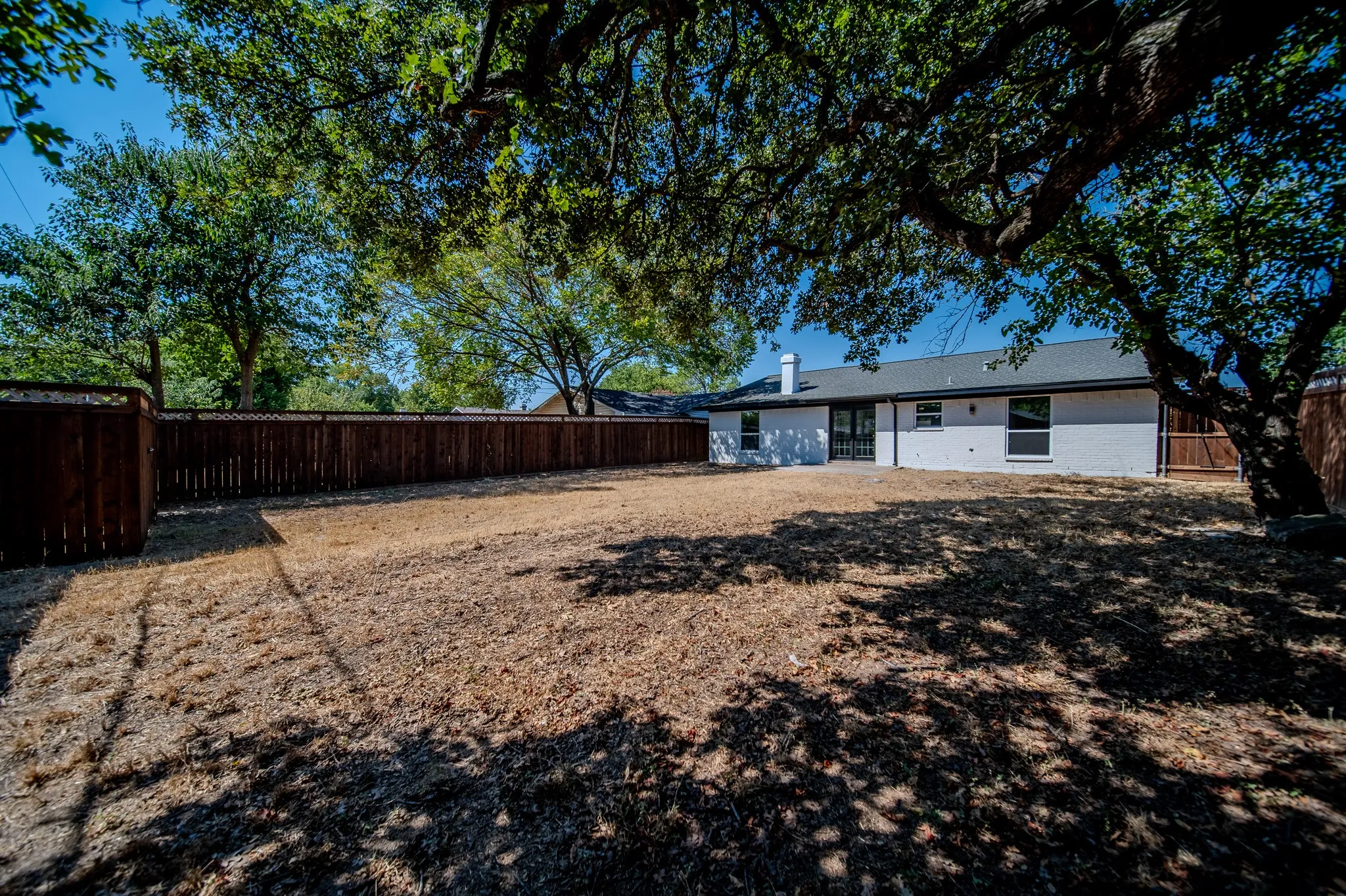 Expansive backyard with a full privacy fence, mature trees providing shade, and a white brick exterior with a dark roof