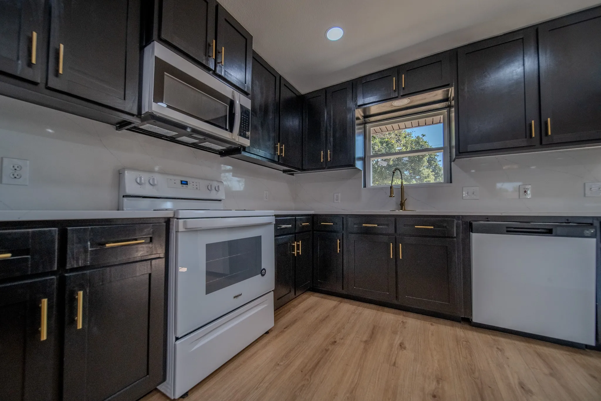 Dark wood-finish cabinetry with brass hardware, white countertops, coordinating white appliances, a stainless steel microwave, and wood-finish flooring