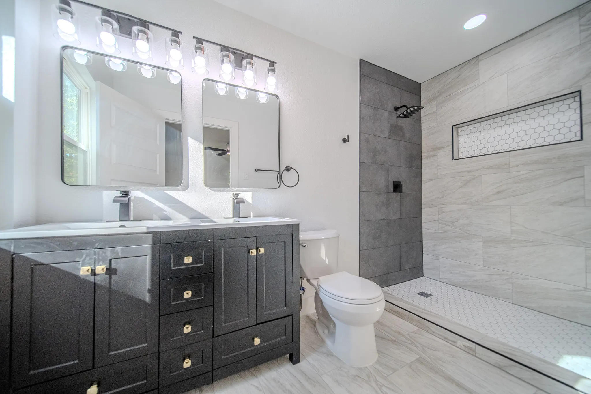Dual vanity with integrated sinks, a walk-in shower featuring a tiled niche, wood-finish flooring, and black fixtures