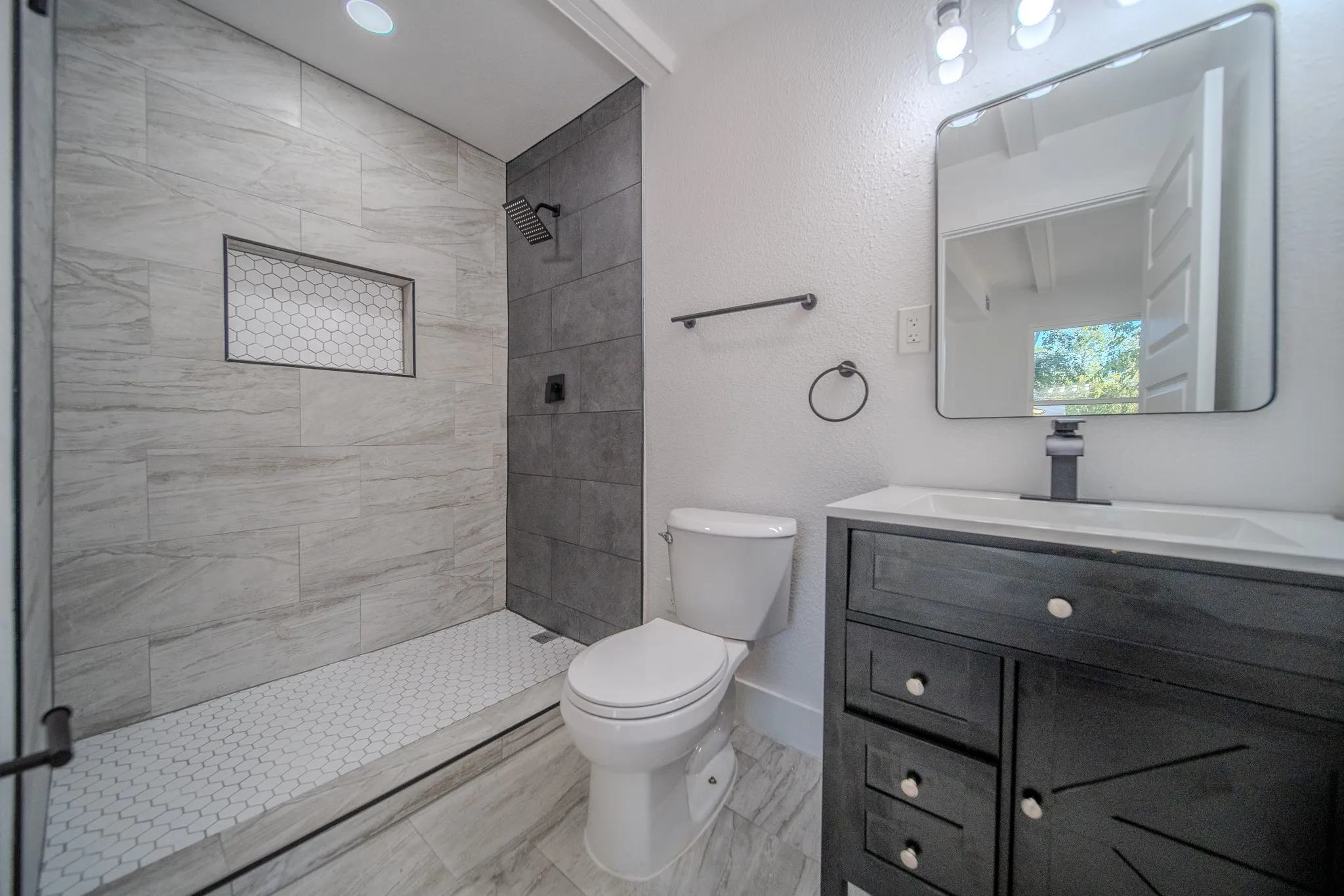 Modern bathroom featuring a walk-in shower with linear tile detailing and frameless glass enclosure, dual-sink vanity with dark cabinetry and quartz countertop, vessel sink with chrome faucet, illuminated mirror with integrated lighting, and porcelain tile flooring throughout