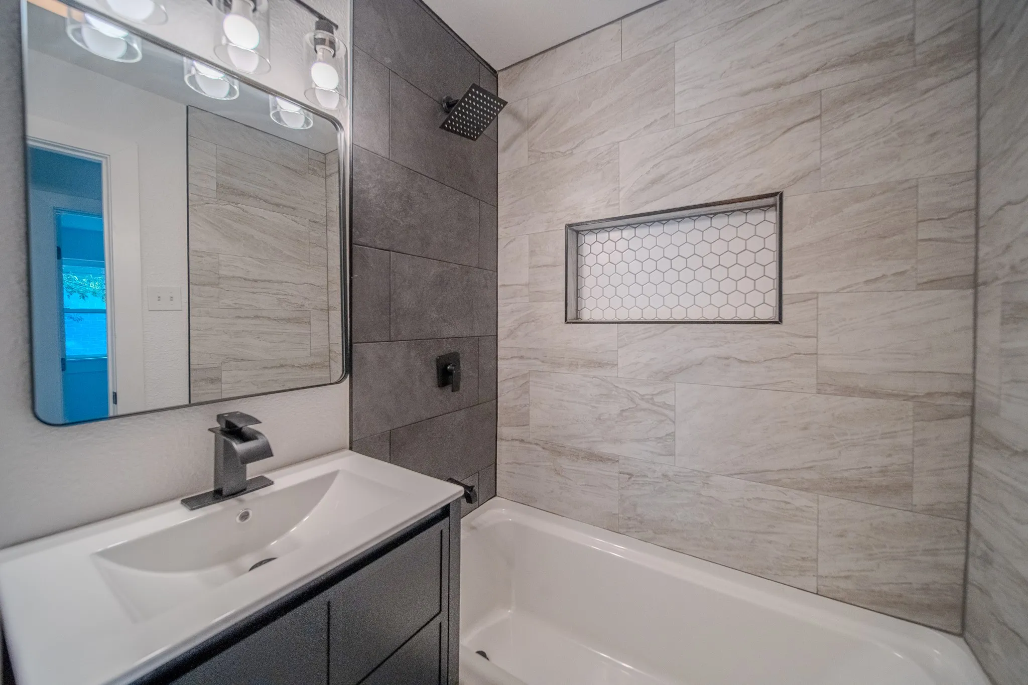 Vanity with integrated sink and matte black faucet, framed mirror with overhead lighting, tiled shower surround with recessed niche, and a contrasting dark tile accent wall