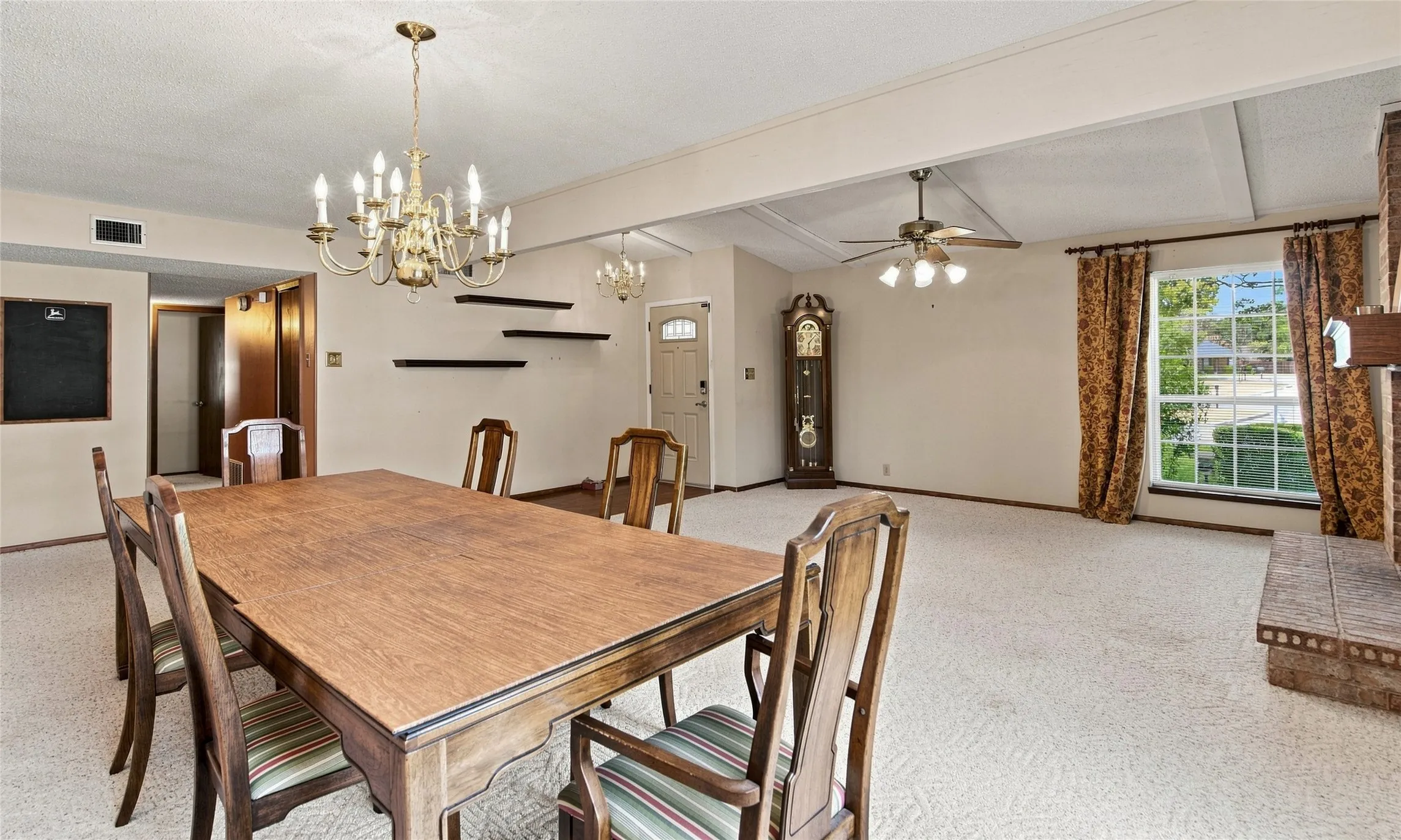 Spacious room featuring a brass chandelier, a ceiling fan, a large window, and exposed ceiling beams