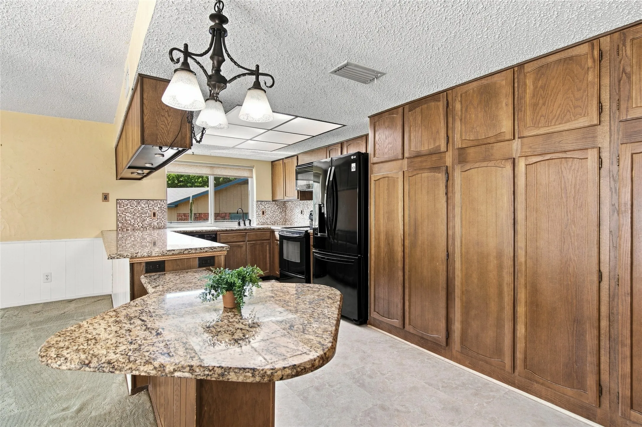 Kitchen featuring extensive wood cabinetry, granite-style countertops, mosaic tile backsplash, black appliances, and white wainscoting
