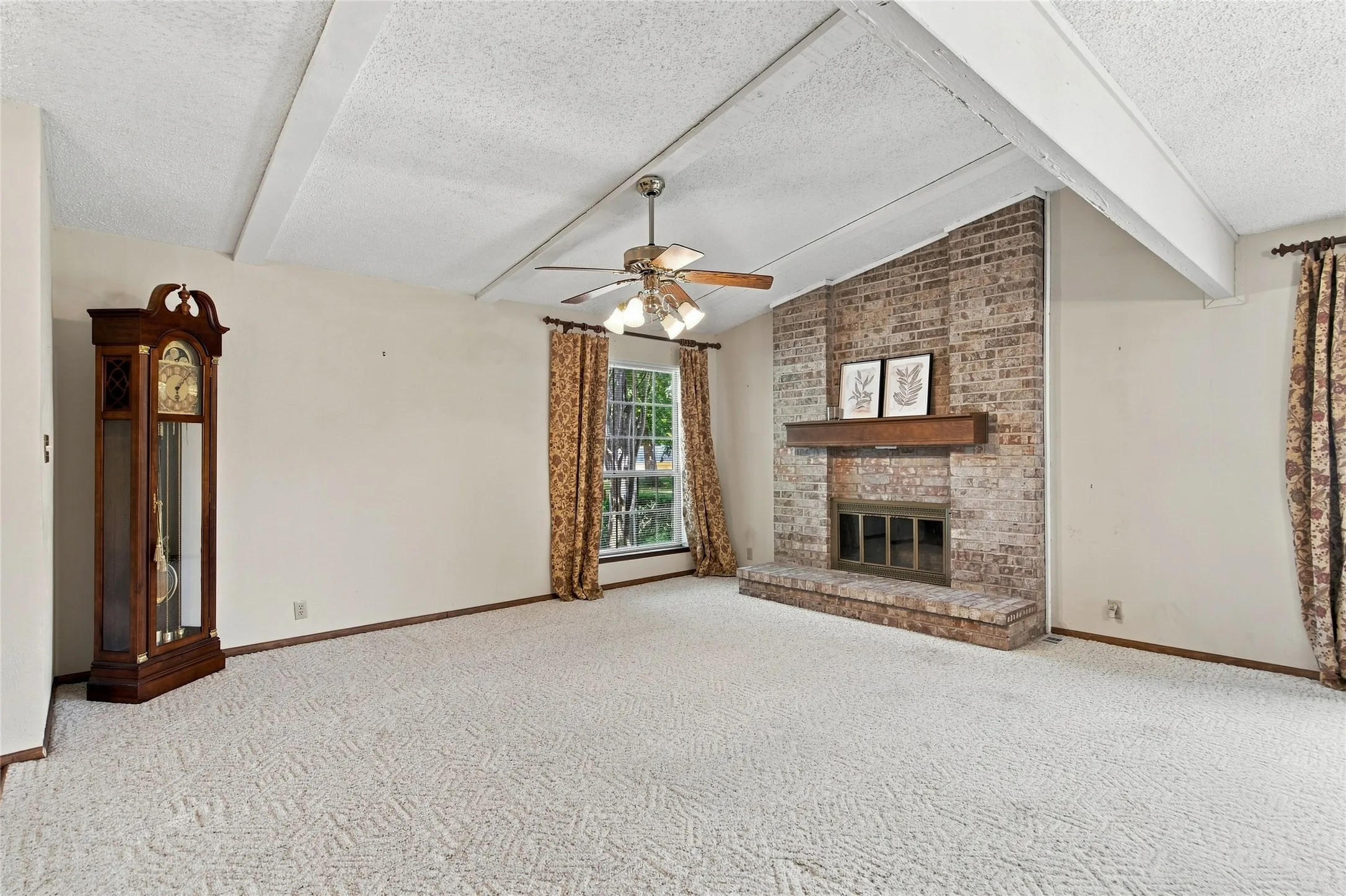 Spacious living area featuring a brick fireplace with a wood mantle, vaulted ceiling with exposed beams, and a ceiling fan