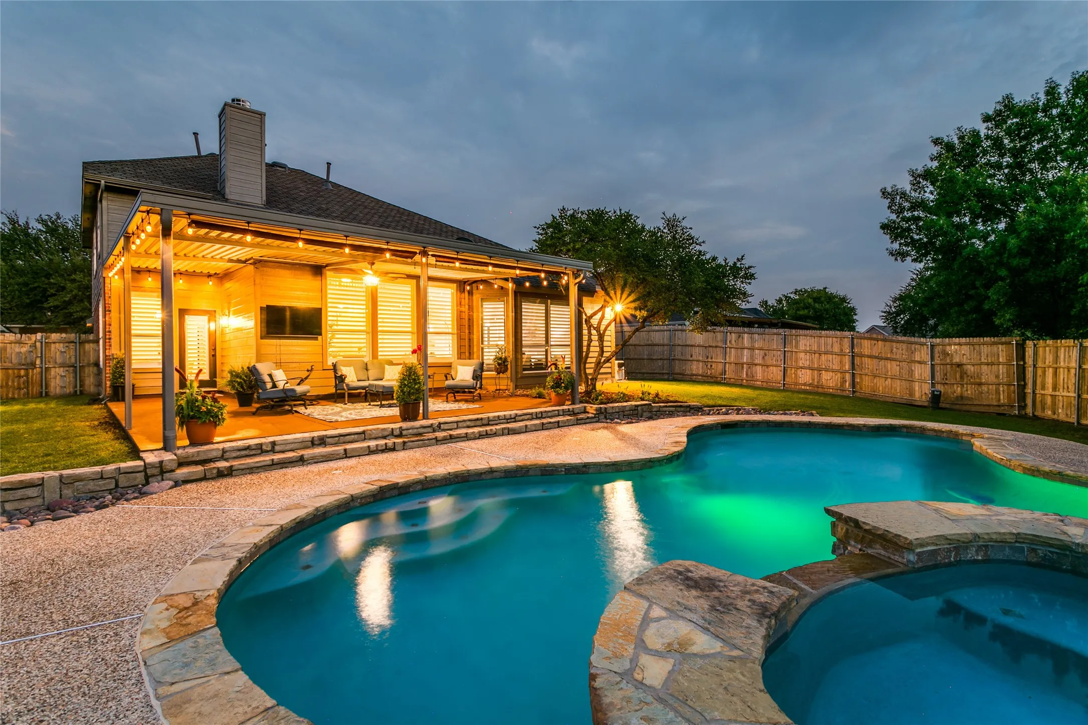 Expansive outdoor living area featuring a covered patio with string lighting, a freeform swimming pool, an integrated spa, and stone coping