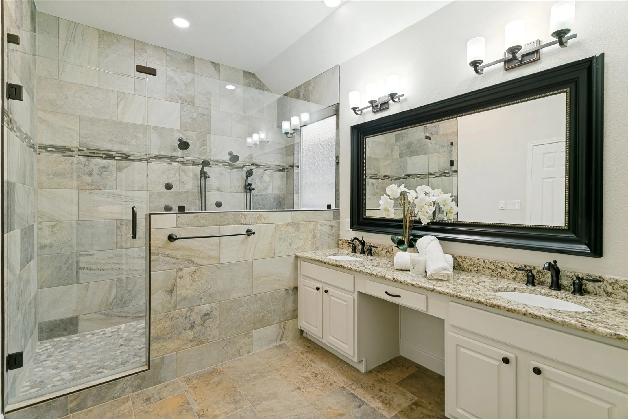 Walk-in shower with tile surround and pebble flooring, dual shower heads, body sprayers. dual vanities with integrated sinks, framed mirror, and tile flooring