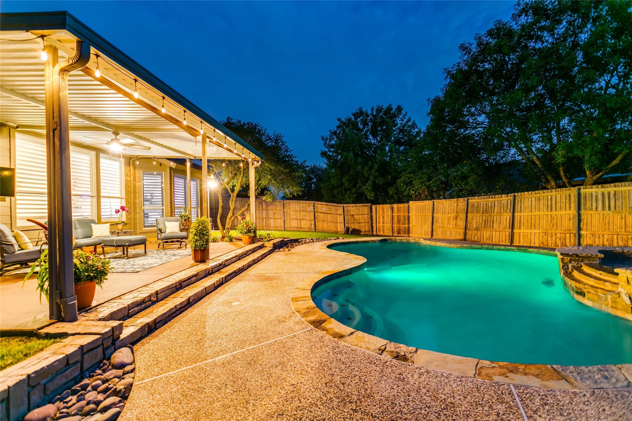 Curved spacious swimming pool with integrated stone spa, expansive covered patio with ceiling fan, wood privacy fencing, and stone retaining wall with integrated steps