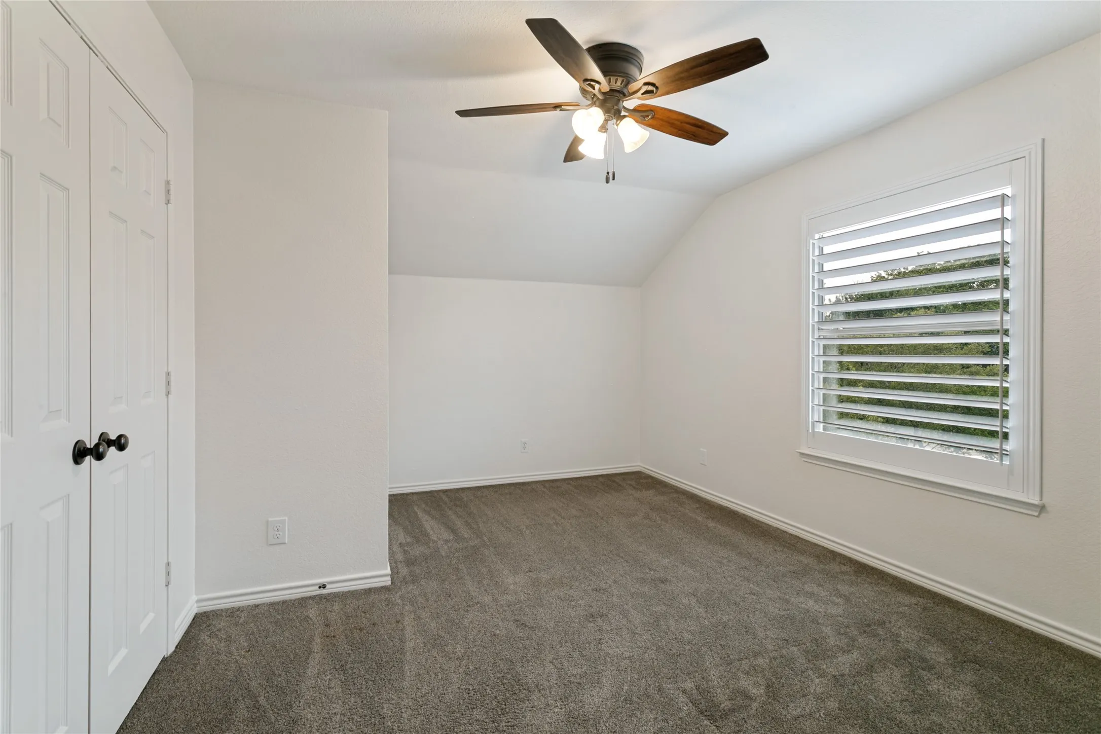 Carpeted room with a ceiling fan, window featuring plantation shutters, and a closet with bifold doors